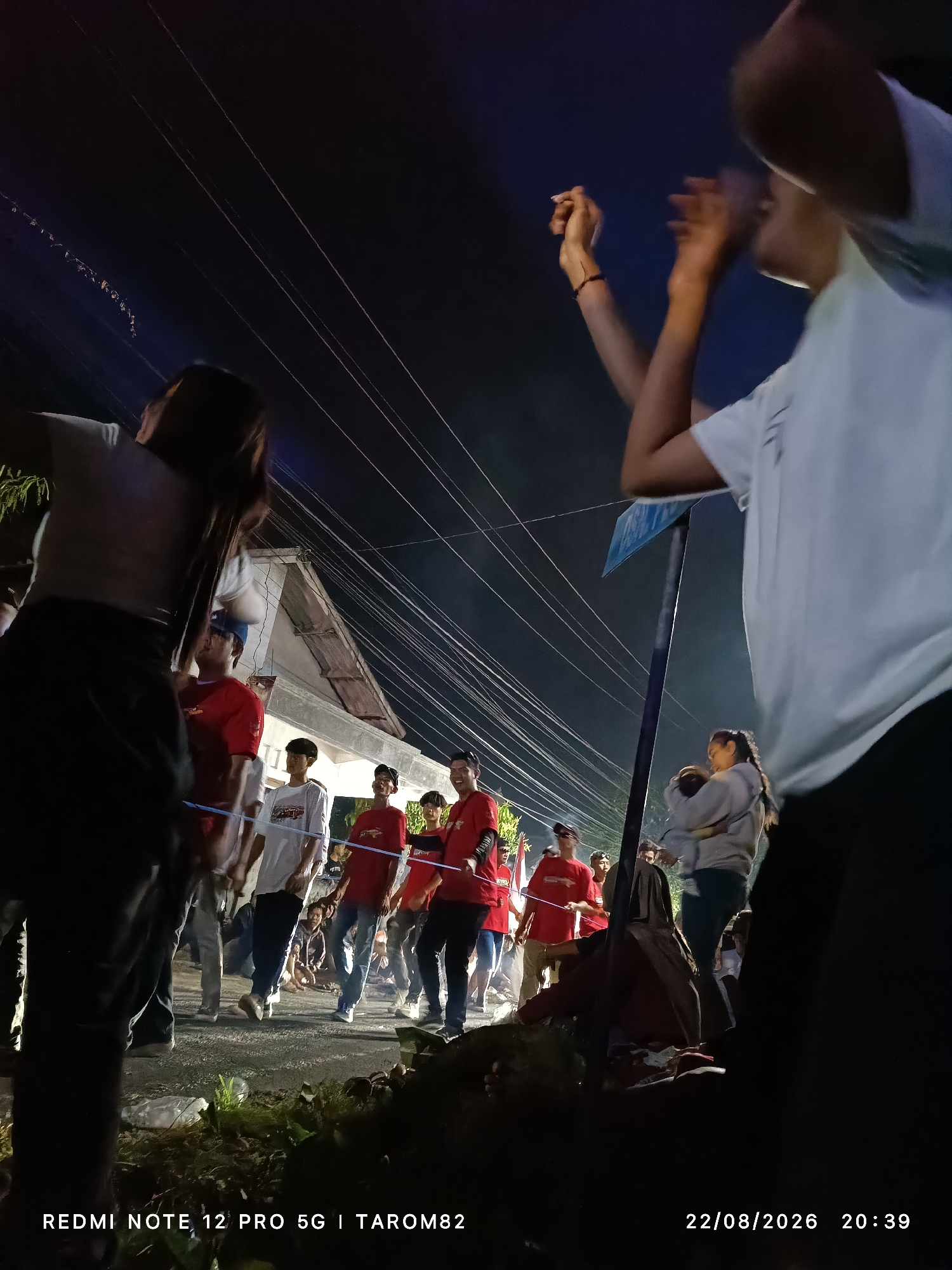 In the city of Mundusewu, Indonesia, on August 22, 2026, at 20:39, a group of young people are gathered outdoors at night. Several individuals are wearing red t-shirts, while others are in white. They appear to be engaged in some form of social activity or event. The background shows a building with a visible roof and numerous power lines stretching across the dark sky. Some people are standing, while others are sitting or crouching on the ground. The overall scene suggests a casual community gathering in a residential area.