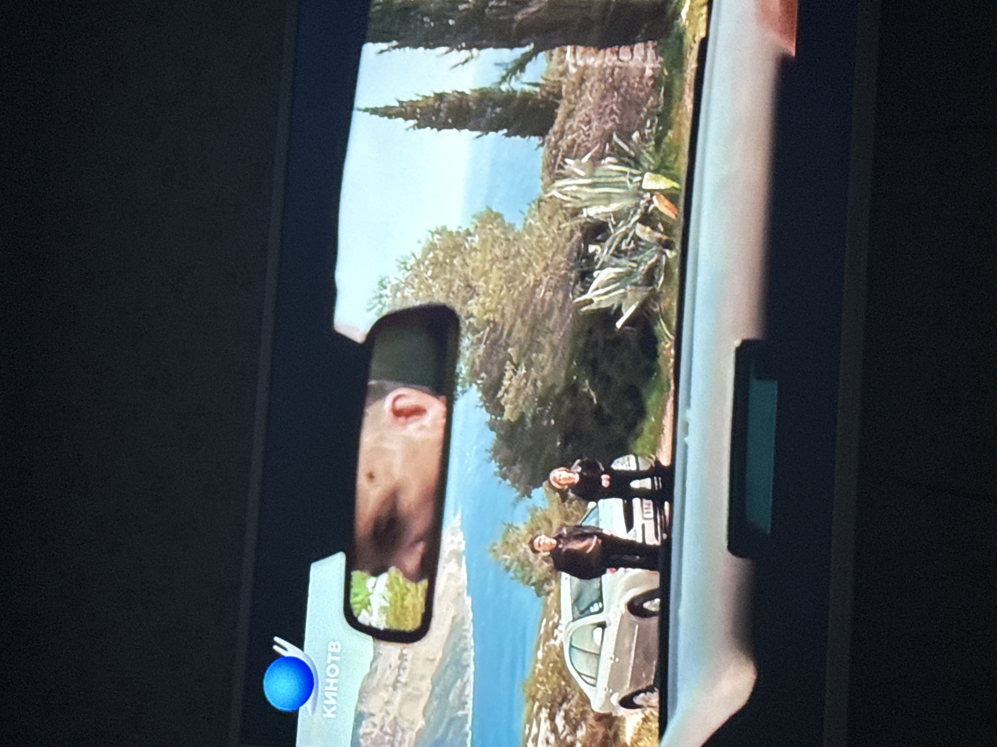 The image depicts a television screen displaying a scene from a movie or show. The scene appears to be filmed from inside a car, looking out through the side window. The side mirror reflects a person's face. Outside the car, two individuals stand near a white vehicle parked on a road or path with a coastal view. The background includes trees, vegetation, and the sea. The location is unknown and there is not enough information to determine if it is in the city of Mytishchi, Russia. A logo "КИНОТВ" is visible on the screen.