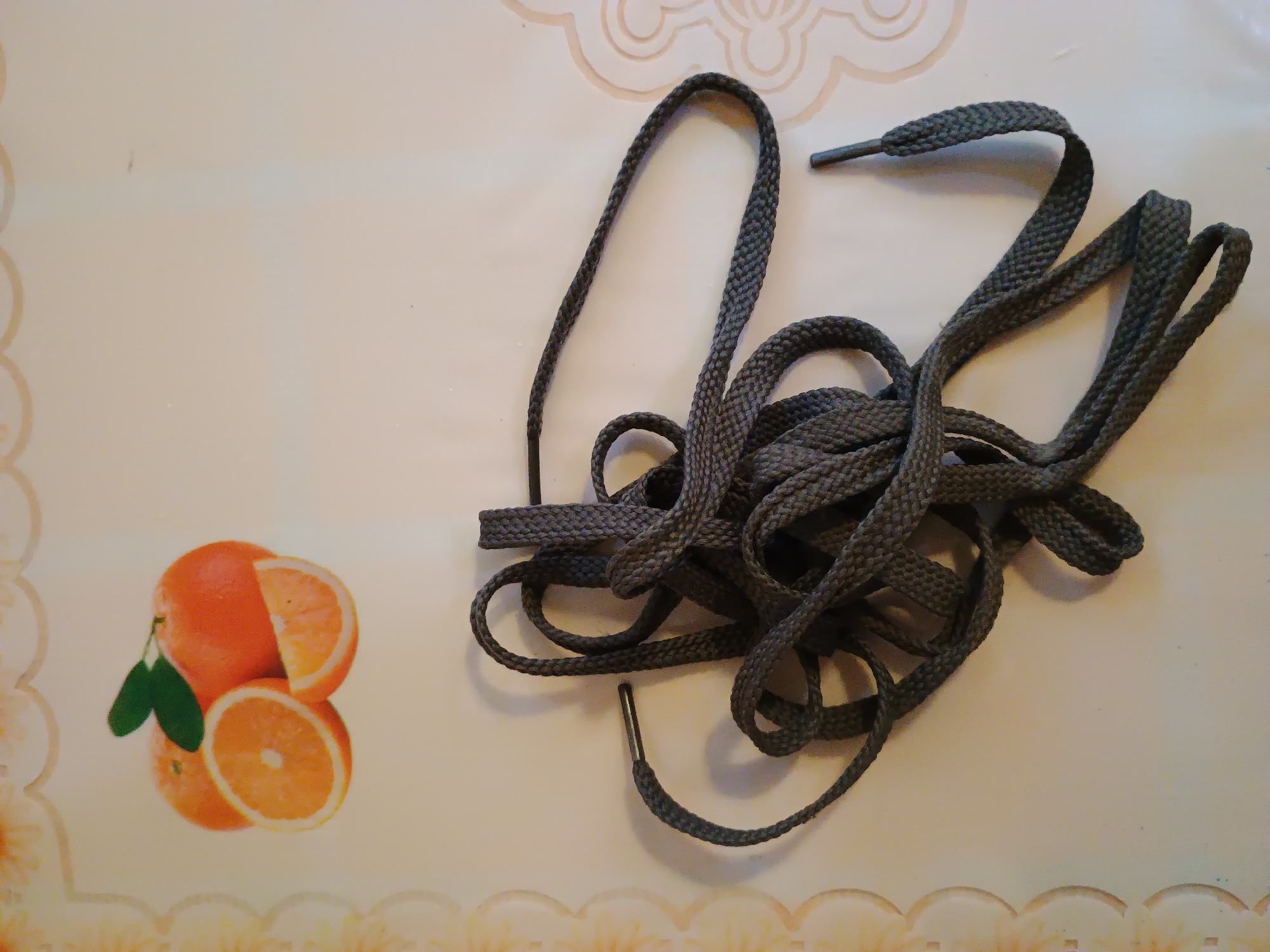 A pile of dark grey shoelaces is arranged on a light-colored surface. The shoelaces are tangled and form loops and coils. The surface appears to be a table or countertop with a decorative border on the left side featuring a pattern of overlapping orange and yellow circles. In the upper left corner of the image, there are three stylized representations of oranges, one whole and two sliced, with green leaves. This scene is indoors. There is no information in the image to suggest a location in the city of Zaporizhzhia, Ukraine.
