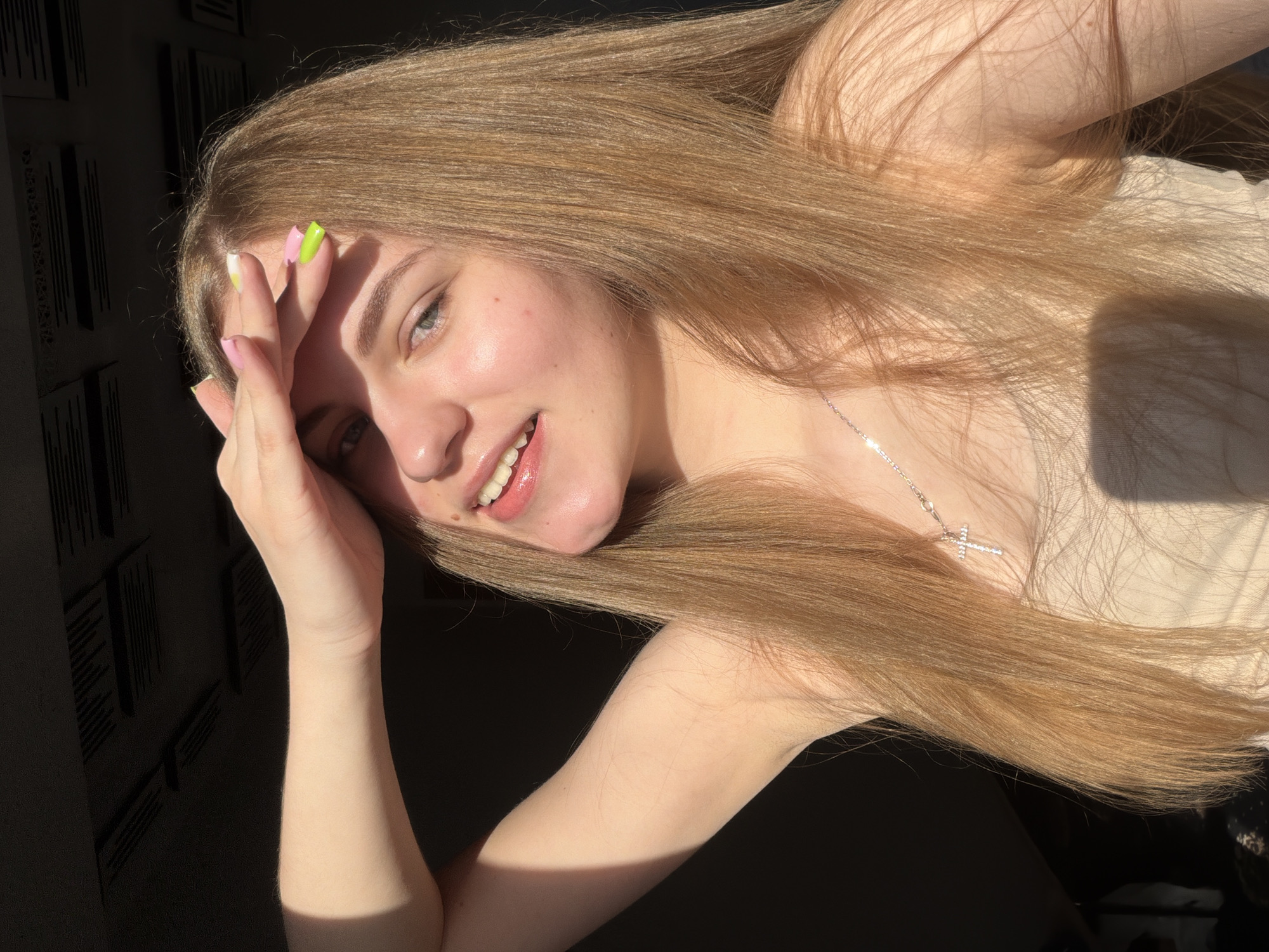 A young woman with long, light brown hair is shown from the chest up in a sunlit indoor environment. She is looking towards the camera with a slight smile and has her right hand raised to her forehead, shielding her eyes from the sun. She is wearing a light-colored tank top and a delicate silver necklace with a cross pendant. Her fingernails are painted with vibrant colors, including pink and neon green. The background is dark, with what appears to be a textured ceiling visible at the top of the frame. The lighting suggests it is daytime, with sunlight casting strong shadows. No specific location in Dnipro, Ukraine can be identified from the image.