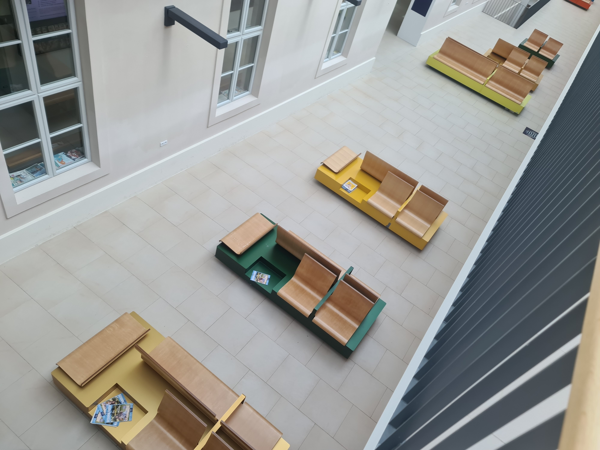The image shows an interior view of a public building, likely a library or exhibition space, with a minimalist design and ample natural light. The flooring consists of large, light-colored square tiles, and the walls are painted a neutral, off-white color. Several modern seating units are strategically placed throughout the space.
In the foreground, a low, yellow seating unit is visible with light wood bench sections. Next to it, another seating unit in a dark green color also features light wood benches. A third seating unit, predominantly yellow, is arranged in a modular fashion, with separate sections of light wood benches. Further back and to the right, a longer, bright green seating unit with multiple light wood bench sections is arranged in a serpentine shape.
The environment appears clean and organized, with no visible people. The architecture includes large windows with white frames, and on one wall, two dark, angular fixtures are mounted. In the windows, some publications or brochures are visible. A dark, slatted wall or partition runs along the right side of the frame, creating a visual division and casting shadows. The overall impression is one of a contemporary, functional, and spacious public area designed for comfortable waiting or reading.
This scene is located in the "Wien Museum MUSA" in Vienna, Austria, specifically in the public area on one of its upper floors, adjacent to the exhibition spaces.