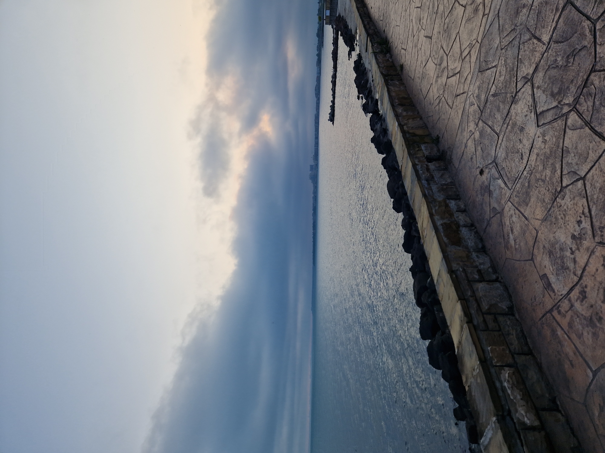 The image depicts a tranquil waterfront scene at twilight, possibly dawn or dusk, in an unknown location. A wide, patterned promenade, appearing to be made of stamped concrete or irregular paving stones in muted brown and grey tones, occupies the lower right portion of the frame, receding into the distance. It is bordered on the left by a low, sturdy stone wall constructed from light-colored, irregularly cut stones. Below this wall, a rugged line of dark, rounded rocks extends into the water, forming a natural or man-made breakwater. The main body of water, possibly a sea, ocean, or large lake, fills the left and central sections, exhibiting a relatively calm surface with subtle ripples. Across the water on the distant horizon, a cityscape or cluster of buildings is faintly discernible. The sky above is largely overcast with expansive grey and bluish-grey clouds, though a soft, warm light, possibly from the sun breaking through, is visible on the right side of the cloud cover. No individuals, animals, or prominent actions are present in the scene.