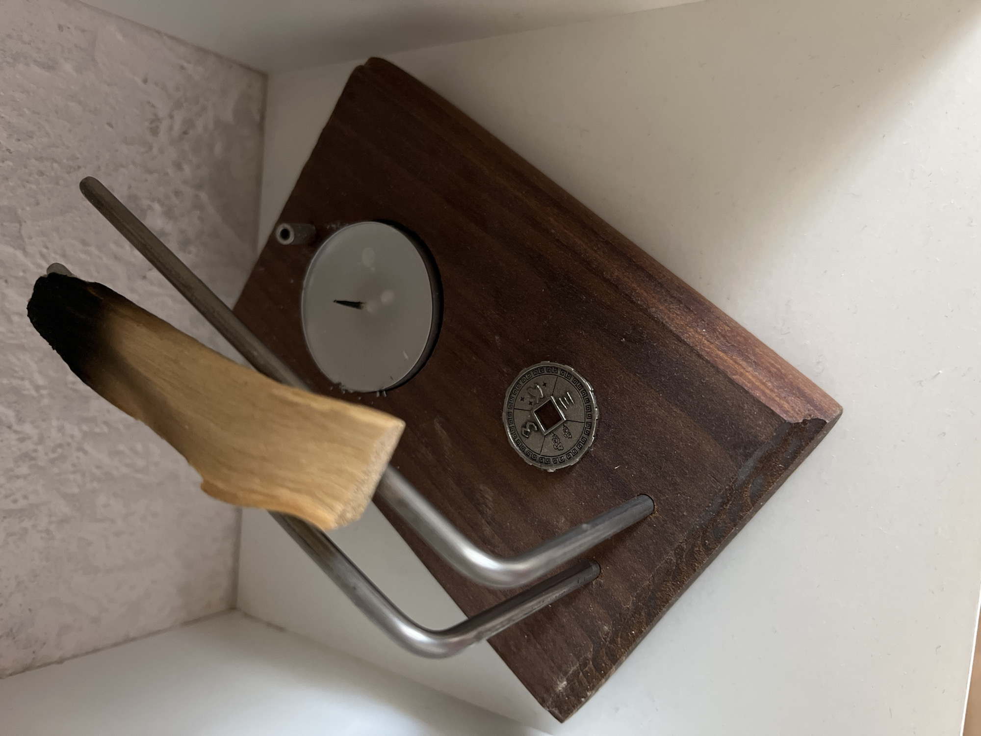 A close-up, eye-level shot displays a spiritual item set against a white, textured background. The item features a dark wooden base, upon which rests a small, silver tea light candle in a metal cup, its wick unlit. To the left of the candle is a decorative metal coin with intricate patterns and a central square hole. A piece of palo santo wood, burnt at one end, is propped upright by two curved metal supports. A thin metal rod extends from behind the palo santo and rests parallel to it. The scene is illuminated by soft, diffused light, casting subtle shadows. The overall impression is one of stillness and ritualistic preparation. There is no indication of Krasnyye Barrikady, Russia.