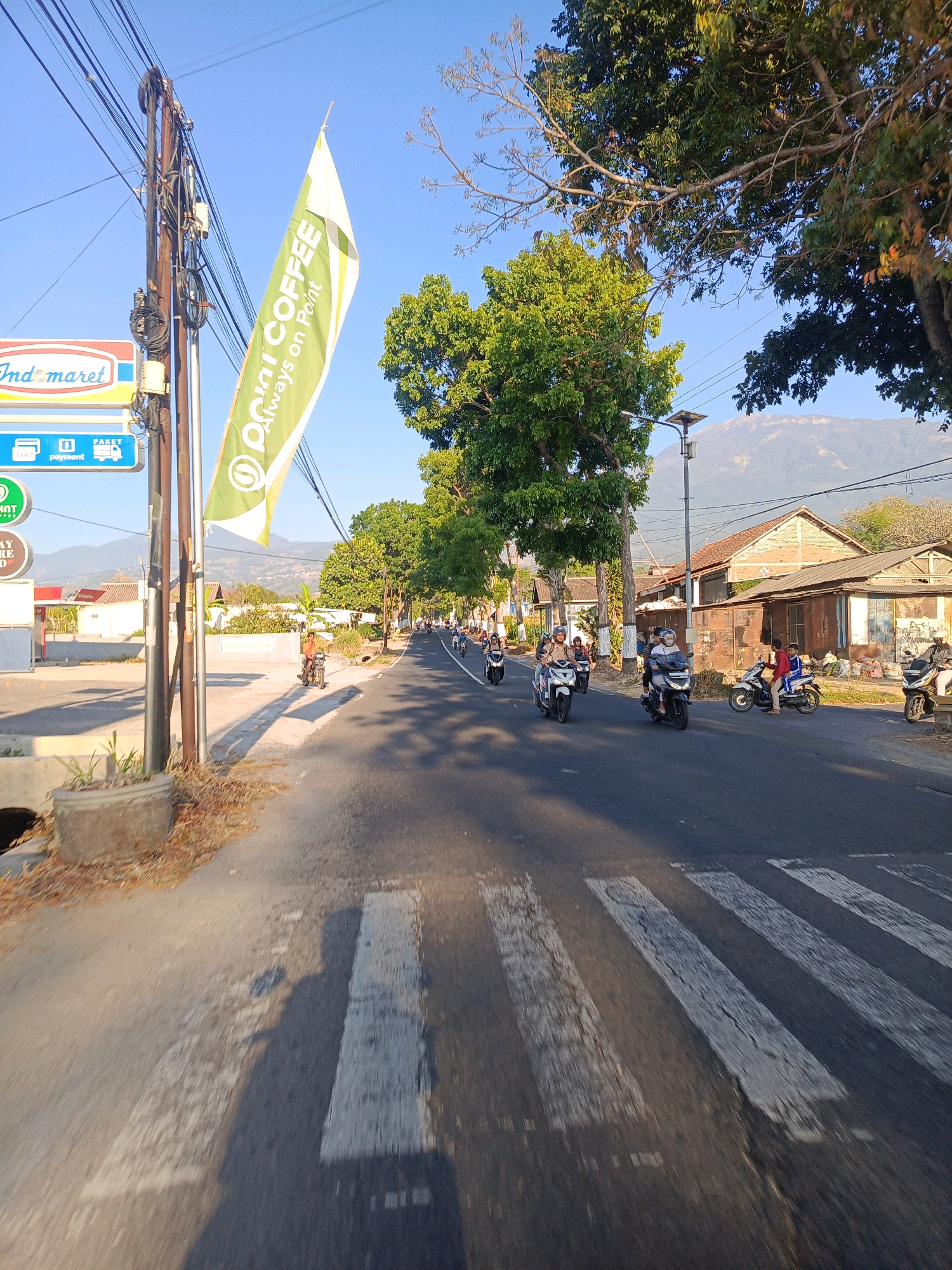 A street scene in Kalang, Indonesia, is captured on a clear, sunny day. The road is paved and shows a pedestrian crossing. Several motorcycles are visible on the road, with riders wearing helmets. On the left side of the road, there is a utility pole with numerous wires, and a green and white flag with text is attached to it. Adjacent to the pole, there are signs for "Indomaret" and what appear to be payment and package delivery services. Trees line the right side of the road, casting shadows. In the background, there are residential buildings with terracotta roofs and a large mountain can be seen in the distance. The perspective suggests the photo was taken from a moving vehicle, possibly a motorcycle, given the motion blur in the foreground.