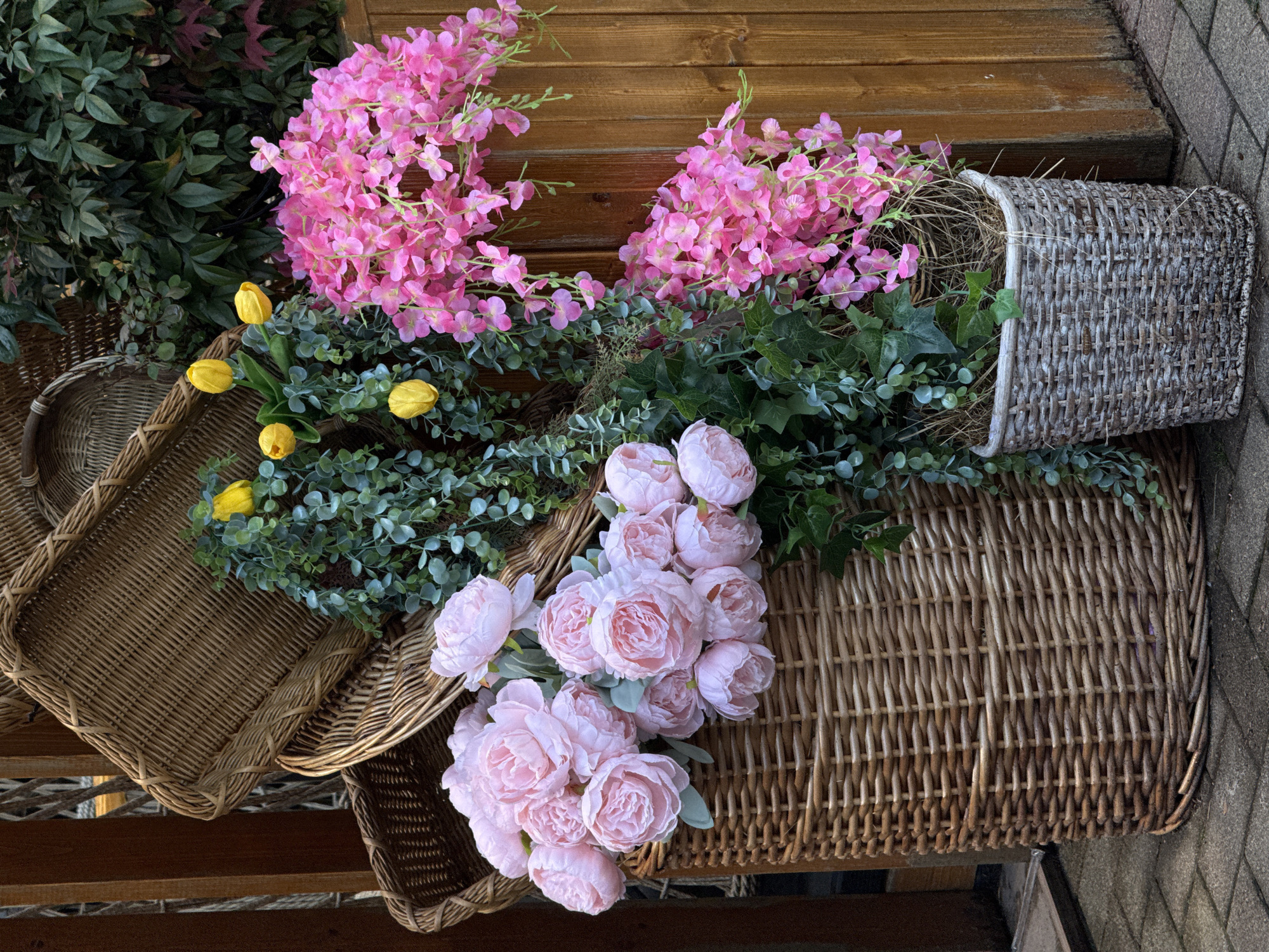 The image presents an arrangement of wicker baskets and artificial flowers on a wooden surface, potentially steps or shelving. There are three main baskets: one square, one rectangular, and one roundish basket tipped on its side. The roundish basket contains pink flowers and greenery. The rectangular basket holds light pink peonies. The square basket contains yellow tulips and greenery. The background includes a wooden wall or structure, suggesting an outdoor setting. The location, Sochi, Russia, is not visually verifiable from the image.