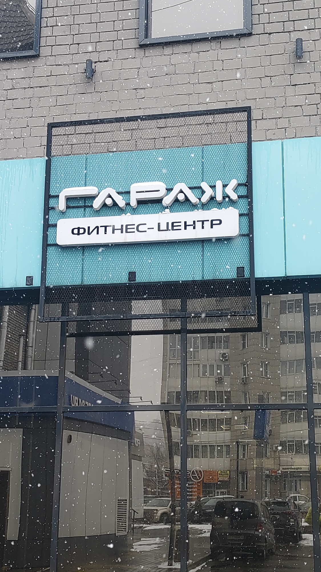 This daytime photograph, taken in Krasnoyarsk, Russia, depicts the exterior of a commercial building on a snowy winter day. Prominent snowflakes are visible throughout the image, falling through the air.
The upper section of the building features a grey brick wall with two dark-framed windows and a cylindrical wall-mounted light fixture. Below the brickwork, a section clad in light blue panels hosts a large sign. The sign, framed in black metal with a black mesh background, displays the name "ГАРАЖ" (GARAGE) in large, stylized white Cyrillic letters. Beneath this, a white rectangular panel bears the inscription "ФИТНЕС-ЦЕНТР" (FITNESS-CENTER) in black Cyrillic letters. The juxtaposition of "Garage" and "Fitness Center" suggests a particular branding or aesthetic for the gym.
The lower half of the image is dominated by a large glass facade, reflecting the urban environment across the street. This reflection reveals a multi-story building with numerous windows, several cars (including a white SUV and darker sedans) parked or moving, and a bare tree trunk. Faintly visible text in the reflection includes "МАГАЗИН МЕБЕЛИ" (FURNITURE STORE) and "ВОДКА", indicating nearby businesses.
No people are clearly visible. The overall scene captures the quiet, cold atmosphere of a snowy day in the city, with the fitness center sign standing out against the winter backdrop.