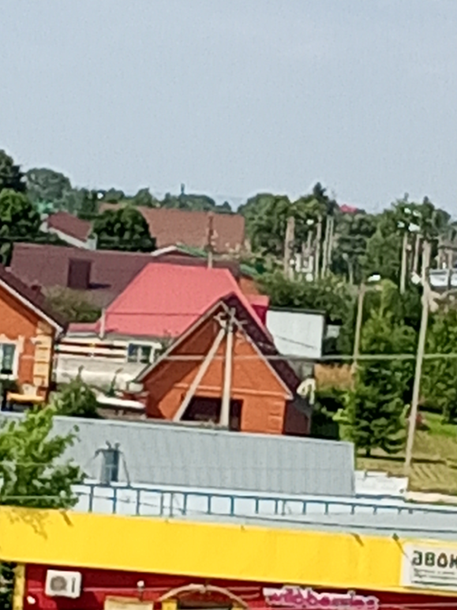 The image depicts a cluster of residential buildings with red and brown tiled roofs, nestled among lush green trees. A prominent red-roofed structure with a distinctive A-frame design is visible in the mid-ground. In the foreground, a bright yellow structure with a red facade is present, featuring signage that includes the word "ЗВОК" in Cyrillic. A white air conditioning unit is mounted on the red facade. The sky is a pale blue. The scene appears to be a residential area within a city. The presence of Cyrillic text suggests a location in Russia, and given the provided location context, it is likely Chistopol.