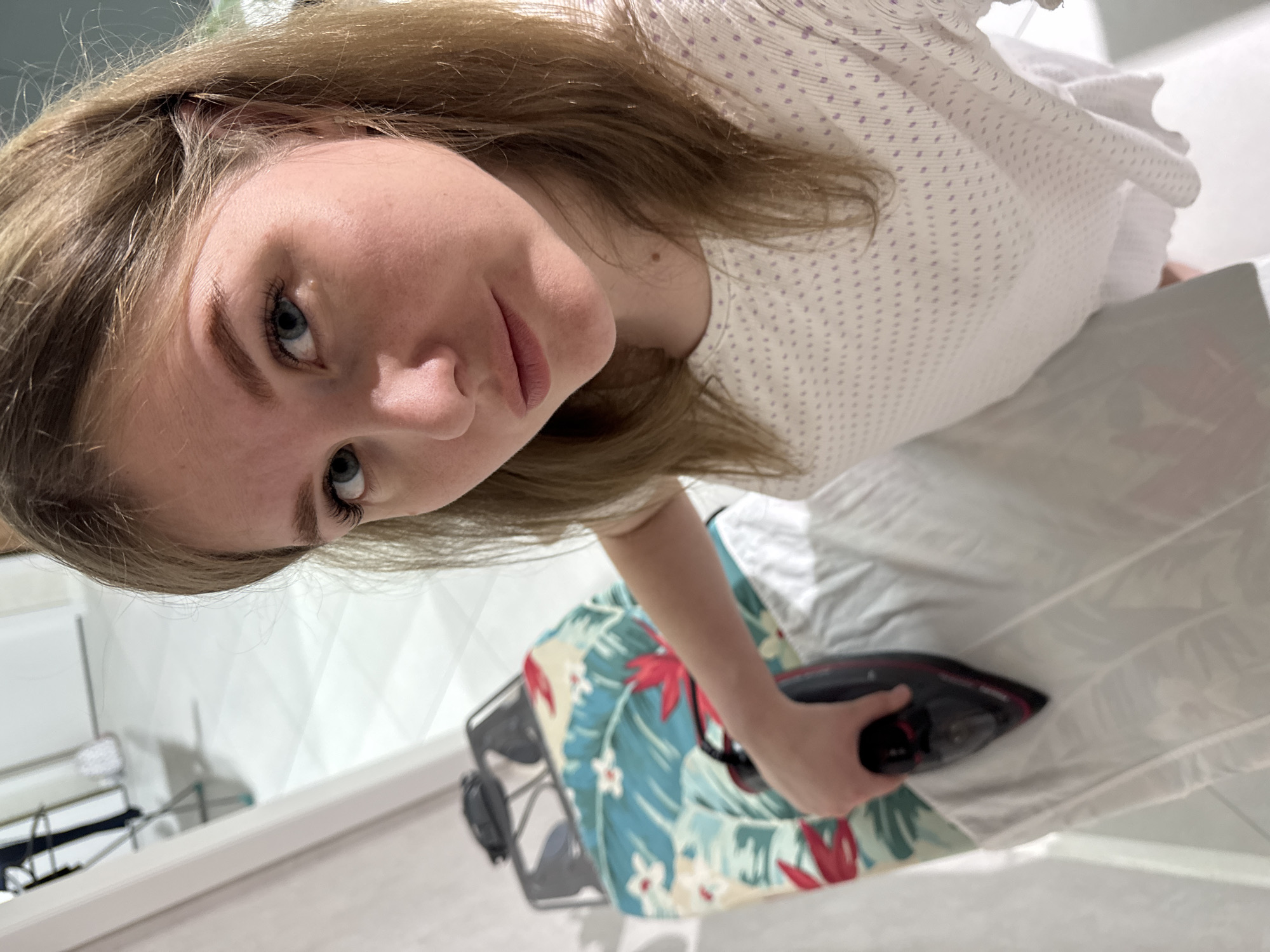 The scene, located indoors in the city of Gornyi (Dvurechenskoe S/P), Russia, features an adult female in the foreground. She has fair skin, light brown hair, and blue eyes, looking directly at the camera with a neutral expression. She wears a white, short-sleeved top with a repeating pattern of small, light purple dots. Her right hand holds a black and red electric iron, which is positioned on a white garment laid across an ironing board. The ironing board is covered with a light blue fabric adorned with a tropical floral pattern of red flowers and green leaves. The background consists of a white tiled floor with a diagonal grid pattern and a white wall or door frame. A blurred structure, possibly a clothes drying rack, is partially visible in the distant left background. The area is brightly and evenly illuminated.