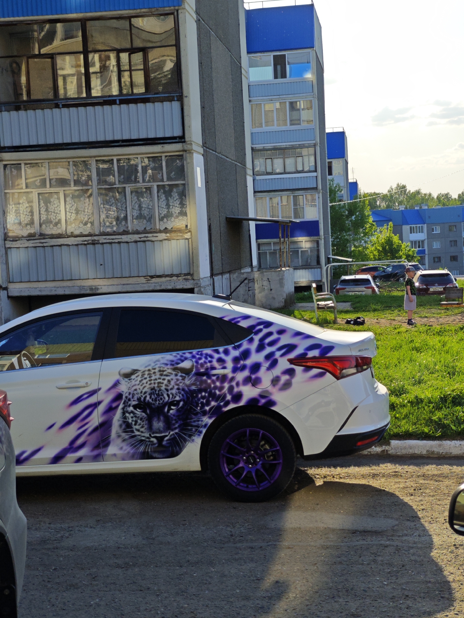 The image captures a scene in Марковский, Russia. A white sedan with a custom purple leopard graphic on its rear passenger side is parked on a paved road next to an apartment building. The building has a combination of grey and white concrete exterior. In the background, there is a grassy area with other parked cars and a young boy standing on the grass. The sky is visible with some clouds.