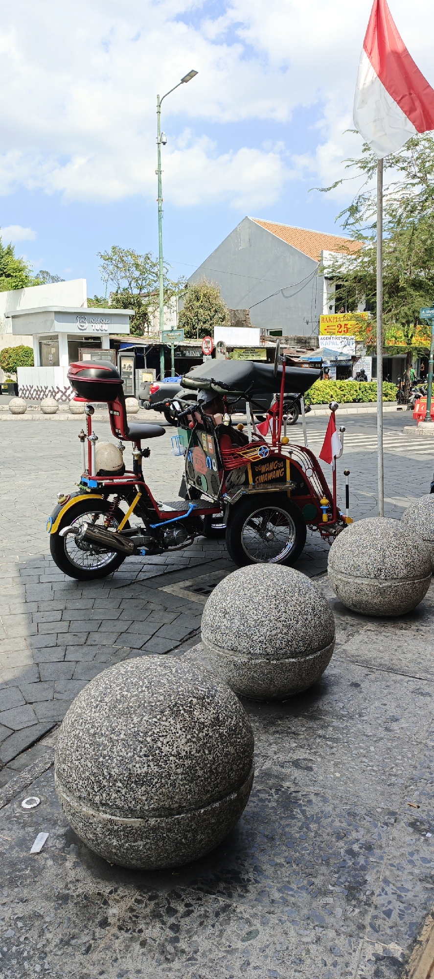 In the city of Yogyakarta, Indonesia, a three-wheeled motor vehicle, painted red and yellow with various colorful decorations, is parked on a cobblestone street. The vehicle, resembling a modified motorcycle with a passenger seat and canopy, displays the text "SINGGAH SINAWANG" on its side. To the right of the vehicle, a large Indonesian flag, red and white, is prominently displayed on a flagpole. In the foreground, three large, spherical stone planters, with a textured gray surface, are positioned on the sidewalk. In the background, a street lamp stands tall, and behind it, buildings with tiled roofs and commercial signage are visible. The sky is blue with scattered clouds, indicating a daytime scene. A small white building with the words "POS POLISI TUGU" and a police emblem is situated to the left of the vehicle.