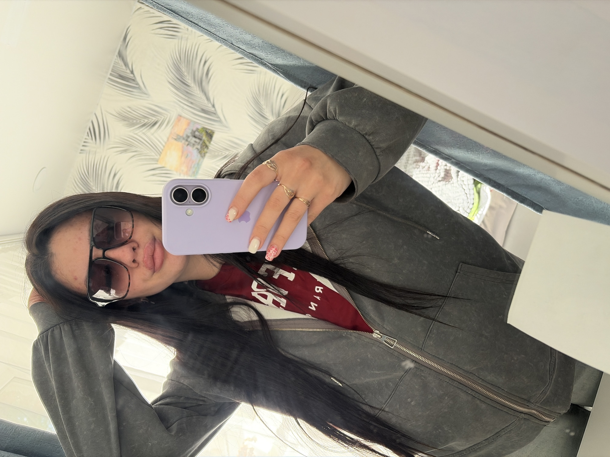 A young woman with long, dark hair is taking a selfie in a mirror. She is wearing sunglasses, a maroon t-shirt, and a gray zip-up hoodie. She is holding a light purple iPhone. The background shows a wall with a white and gray leaf pattern wallpaper and a piece of artwork. The photo is taken indoors, and the location in the city of Фоки, Russia is not discernible from the image.
