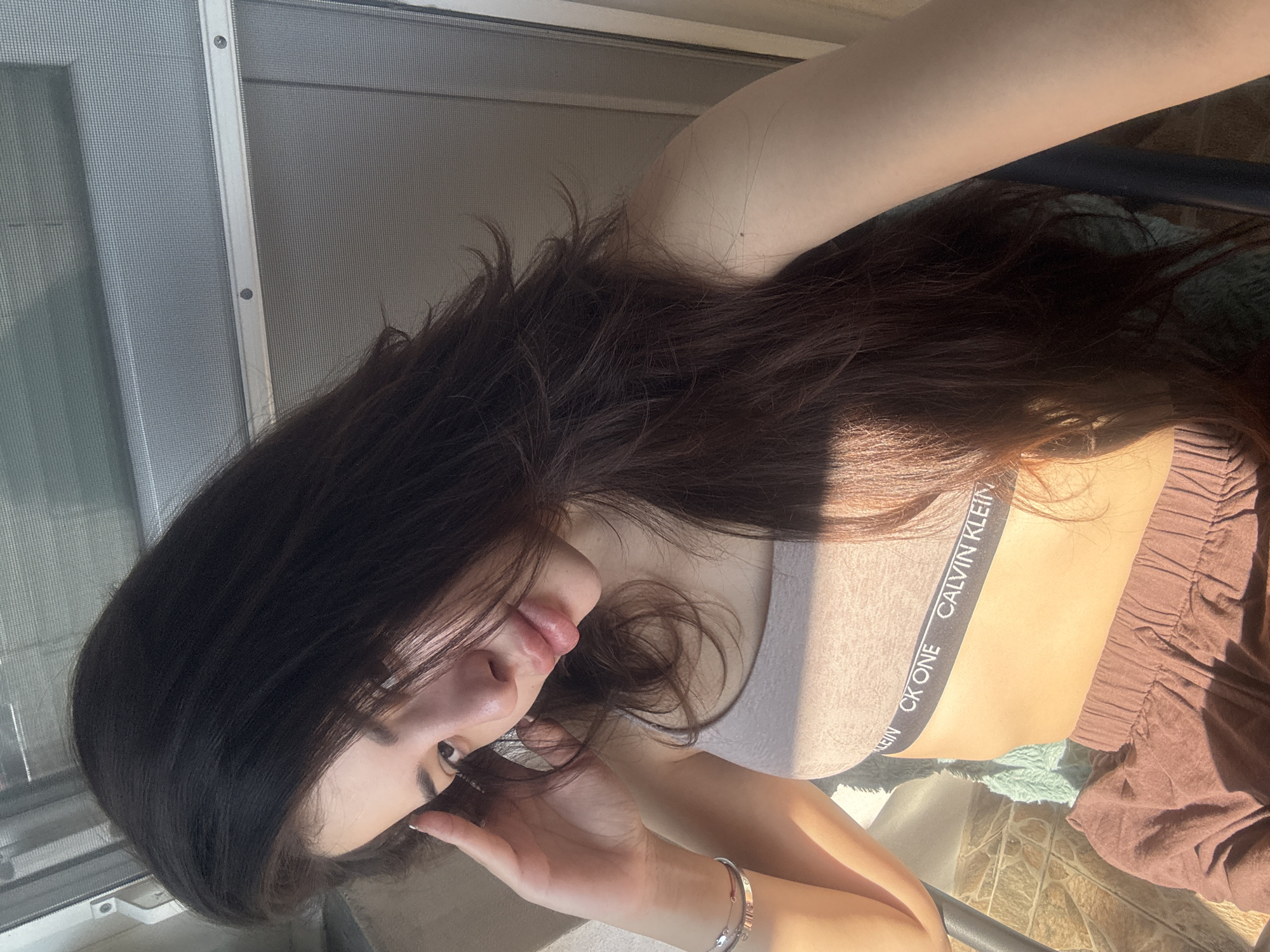 A young female subject is depicted in a close-up, selfie-style photograph, illuminated by strong natural sunlight entering from the left. The scene is located in Plovdiv, Bulgaria. She has long, dark brown hair, styled with layers, which falls over her shoulders and chest. Her left hand is raised, with fingers touching her temple and forehead area, partially obscuring the upper part of her left eye. She is wearing a light beige or pale pink Calvin Klein "CK ONE" sports bra, clearly visible with the brand name printed in black on the elastic band. Her lower garment consists of loose-fitting, dark reddish-brown shorts or loungewear bottoms. On her left wrist, she wears two bracelets: one silver-toned bangle and another thinner bracelet with a red band and a small metallic charm. Her nails have a light-colored polish. She is seated, likely in a dark-framed chair, with a light-colored, possibly textured fabric visible beneath her. To her right and behind her, a window or door with a grey mesh screen and a metallic frame with visible screws is present. To her lower left, a section of tiled flooring, patterned in light and dark brown tones, is visible, alongside a light blue-green textured fabric item.