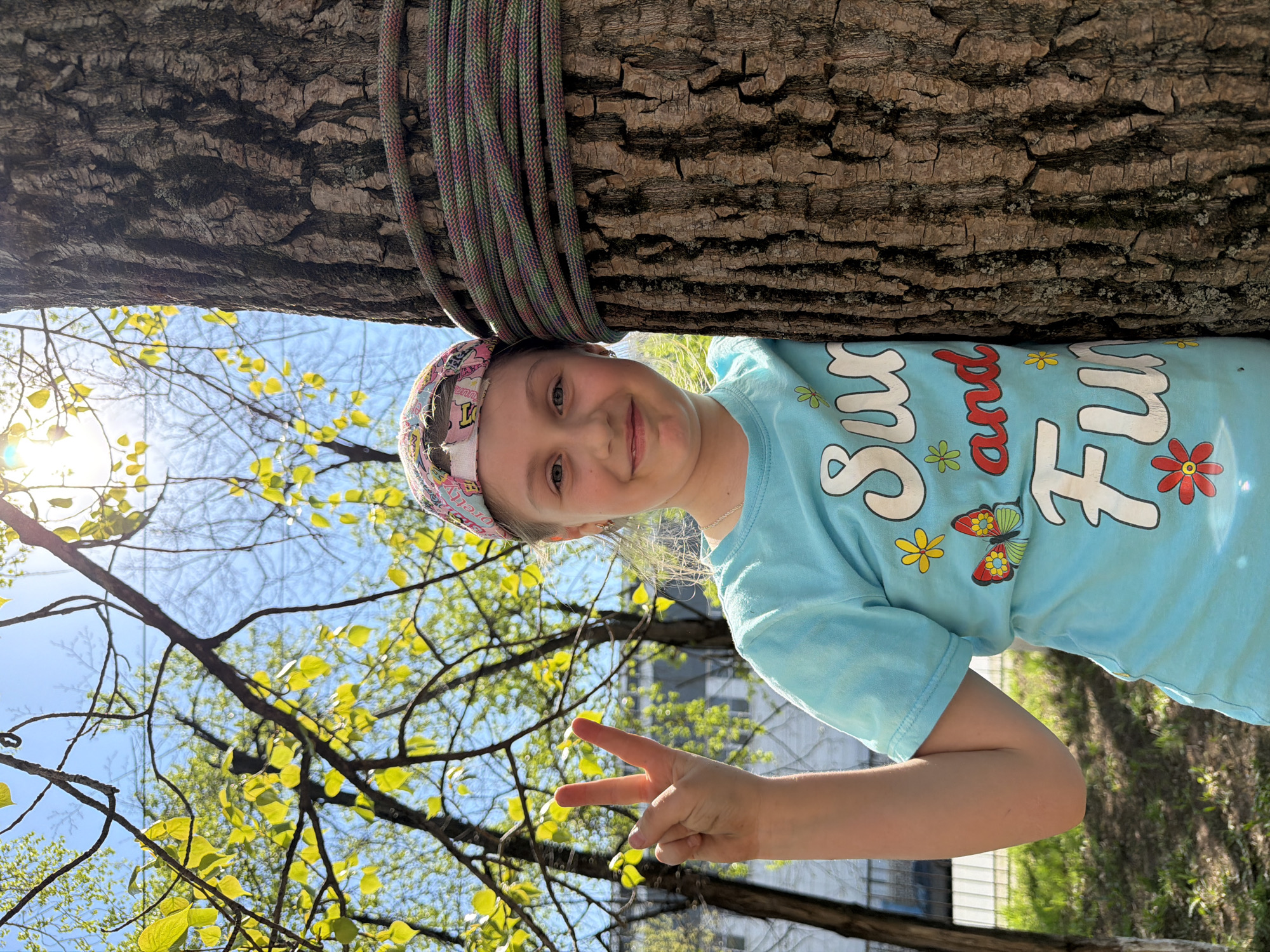 A young girl stands next to a tree with a rope coiled around it. She is smiling and holding up a peace sign with her right hand. She wears a blue t-shirt with a colorful design and a patterned headband. The background includes branches with green leaves and a bright blue sky. The location cannot be determined to be Izhevsk, Russia.
