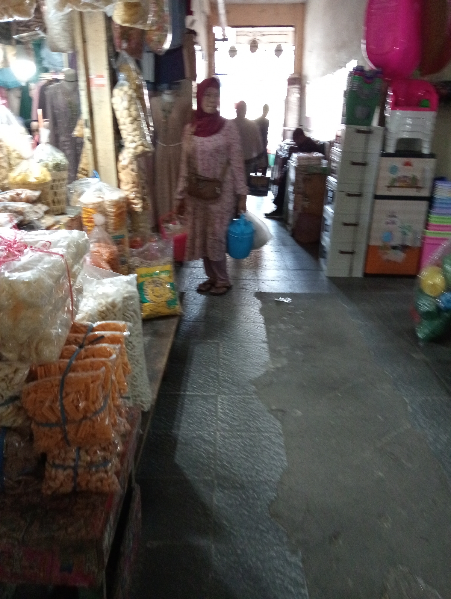 A blurry, indoor scene depicts a market stall in Grabag, Indonesia. A woman wearing a pink hijab and a patterned dress is visible, carrying a blue bucket. To her left, various packaged snacks are stacked on shelves and a table, including items in clear plastic bags and some in orange packaging tied with blue string. In the background, stacked plastic containers and large pink balloons are also present, suggesting a retail environment. The floor appears to be made of dark stone tiles.