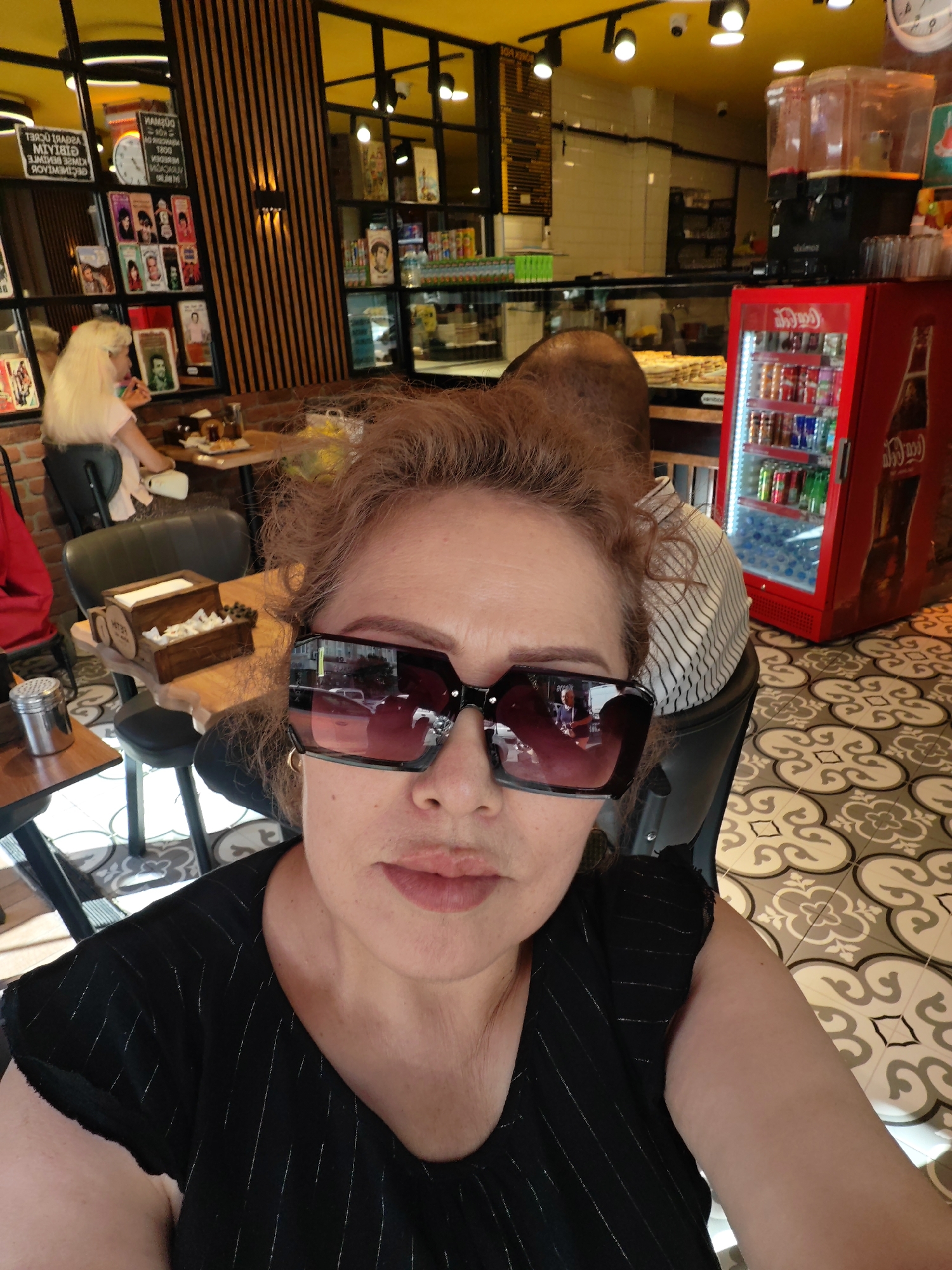 The image shows a close-up of a woman wearing large, dark-rimmed sunglasses and a black top with subtle silver pinstripes. She has reddish-brown, curly hair and is looking directly at the camera. The background suggests an indoor setting, possibly a cafe or restaurant. To the left, a woman with blonde hair is seated at a table, and a red Coca-Cola branded refrigerator is visible on the right. The floor has a patterned tile design. There are also shelves filled with various items, including what appear to be books or magazines. The overall impression is that of a candid snapshot taken in a public establishment. There is no indication of the specific location within Istanbul, Turkey.