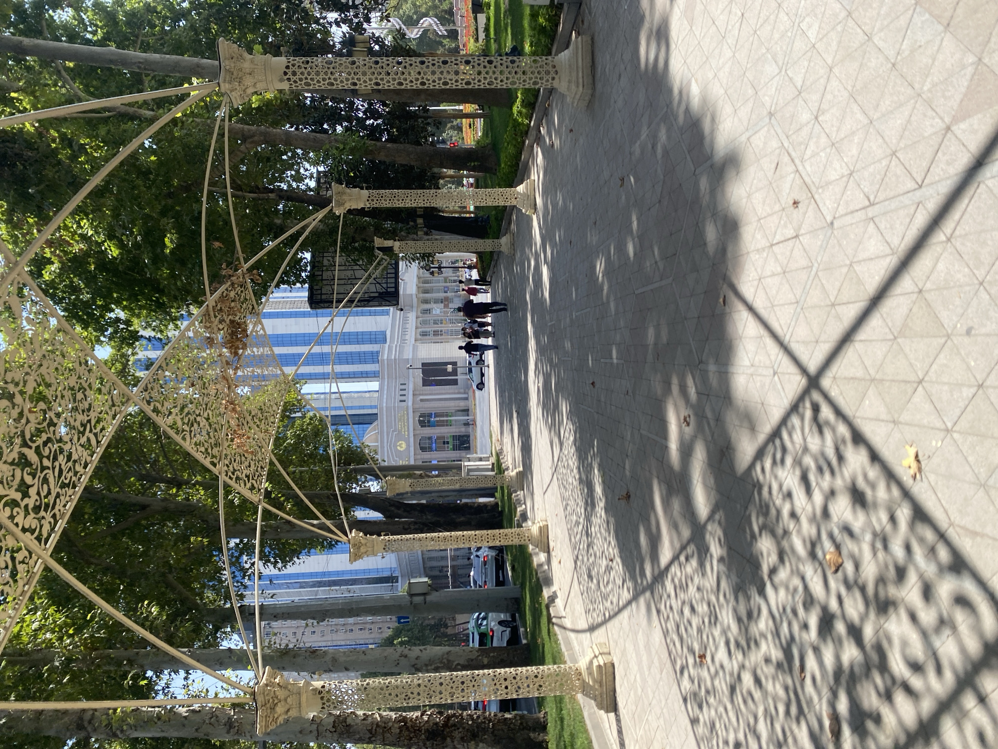 The image depicts a pedestrian walkway in Dushanbe, Tajikistan, leading towards a grand building with a blue, modern facade. The walkway is paved with light-colored tiles and is covered by a decorative white metal canopy supported by ornate pillars. Trees with lush green foliage line the walkway on both sides, casting dappled shadows on the ground. Several people are walking along the path, dressed in casual attire. In the background, vehicles are visible on a street adjacent to the walkway, and a grassy area with flower beds can be seen to the right. The overall scene suggests a well-maintained public space on a bright, sunny day.