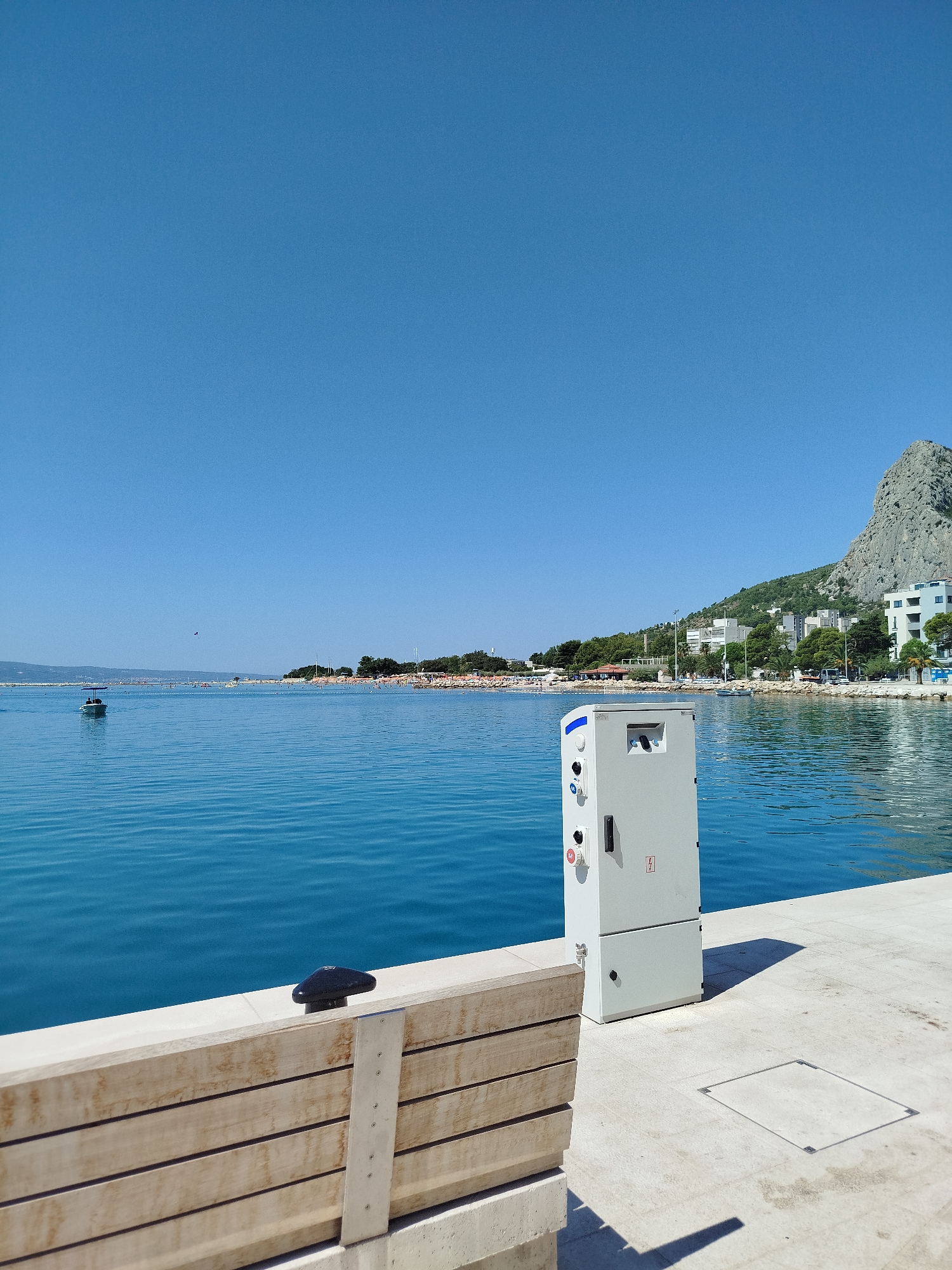 This is a bright, sunny daytime photograph taken from a waterfront promenade in Omiš, Croatia, looking out over the Adriatic Sea. The sky is a clear, vibrant blue, with no clouds visible. The sea is a deep, calm turquoise, with gentle ripples on the surface.
In the foreground, to the right, is a light grey utility box with various electrical outlets and indicator lights. To the left of the utility box, and partially in the foreground, is a wooden bench with concrete supports. A black, mushroom-shaped cleat is visible on the edge of the promenade behind the bench. The promenade itself is paved with light-colored square tiles.
In the middle ground, on the water, a small white boat with a blue canopy is moving away from the shore. Further out in the sea, a long, crowded beach is visible, lined with umbrellas and dotted with people. A thin strip of green trees and buildings is present along the coastline beyond the beach.
In the background, a large, rocky mountain rises steeply from the land. Several buildings, some appearing to be apartment blocks, are nestled on the lower slopes of the mountain. The overall impression is one of a lively coastal town on a beautiful summer day.