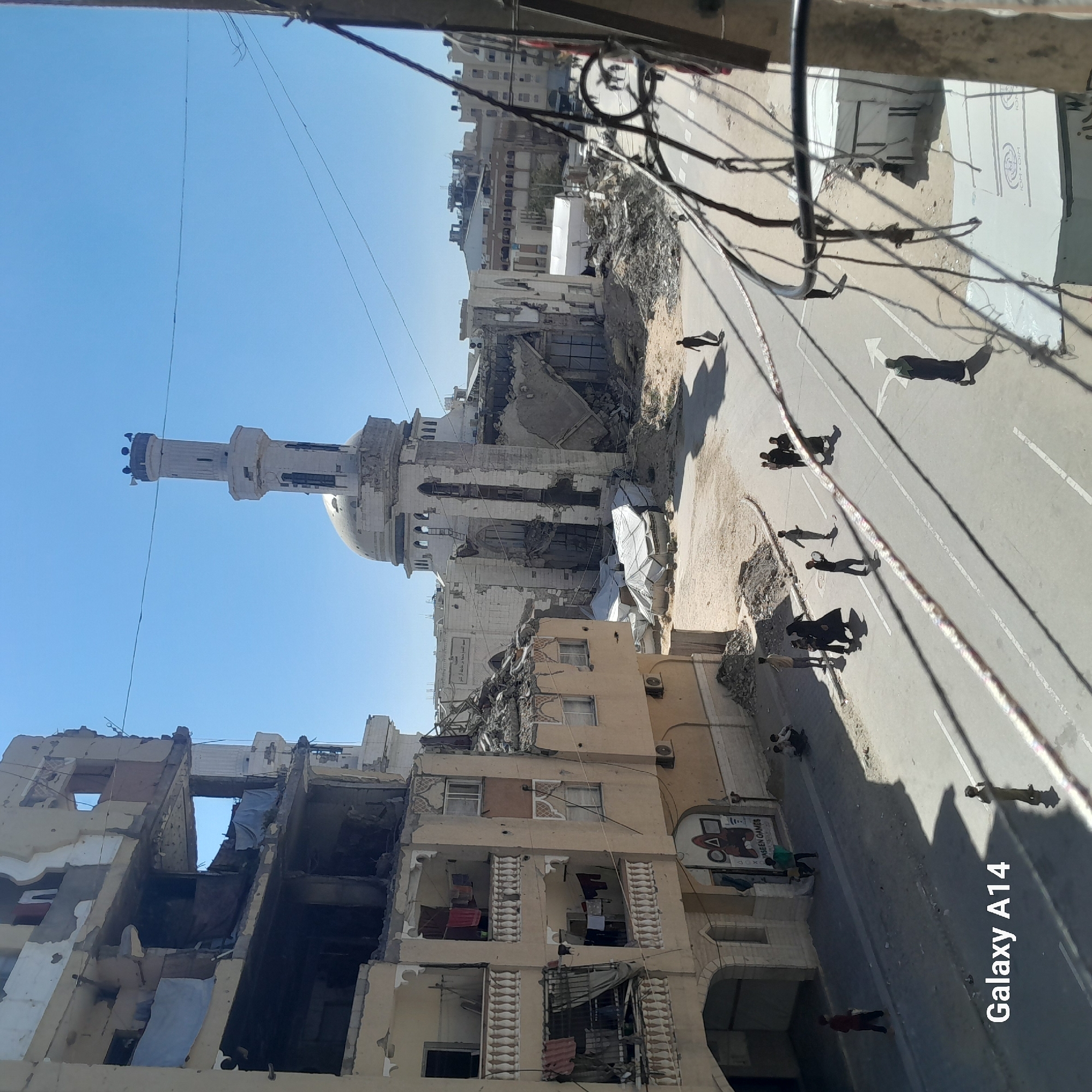 This media file captures a wide-angle view of a heavily damaged urban street scene under a clear, bright blue sky. On the left, multi-story buildings are in various states of ruin, with walls missing, exposed interiors, and rubble piled at their bases. One building with ornate window frames shows severe damage to its upper floors, revealing rooms. Air conditioning units are visible on building exteriors.
In the mid-ground, a prominent mosque with a large dome and a tall minaret stands amidst the destruction. While showing signs of damage, it appears more structurally intact than many surrounding buildings. White tarpaulins or temporary shelters are visible near the mosque, possibly covering damaged areas or serving as temporary structures.
The paved street in the foreground features white lane markings, including a directional arrow. Several individuals, casting long, distinct shadows, are walking or standing along the street and pavement. Their elongated shadows suggest it is either early morning or late afternoon. Large amounts of debris and rubble are scattered along the roadside and among the buildings. Overhead, numerous utility wires crisscross the sky, some appearing tangled.
Visible text includes "YASEEN GAMES" on a sign near a building, along with Arabic script. Another tarp displays partial text, including "FOMOIM" and "ION," potentially related to an organization like IOM. The watermark "Galaxy A14" is present in the bottom right corner of the image. The overall scene depicts a landscape of significant devastation, likely from conflict or disaster, with daily life seemingly continuing amidst the ruins.