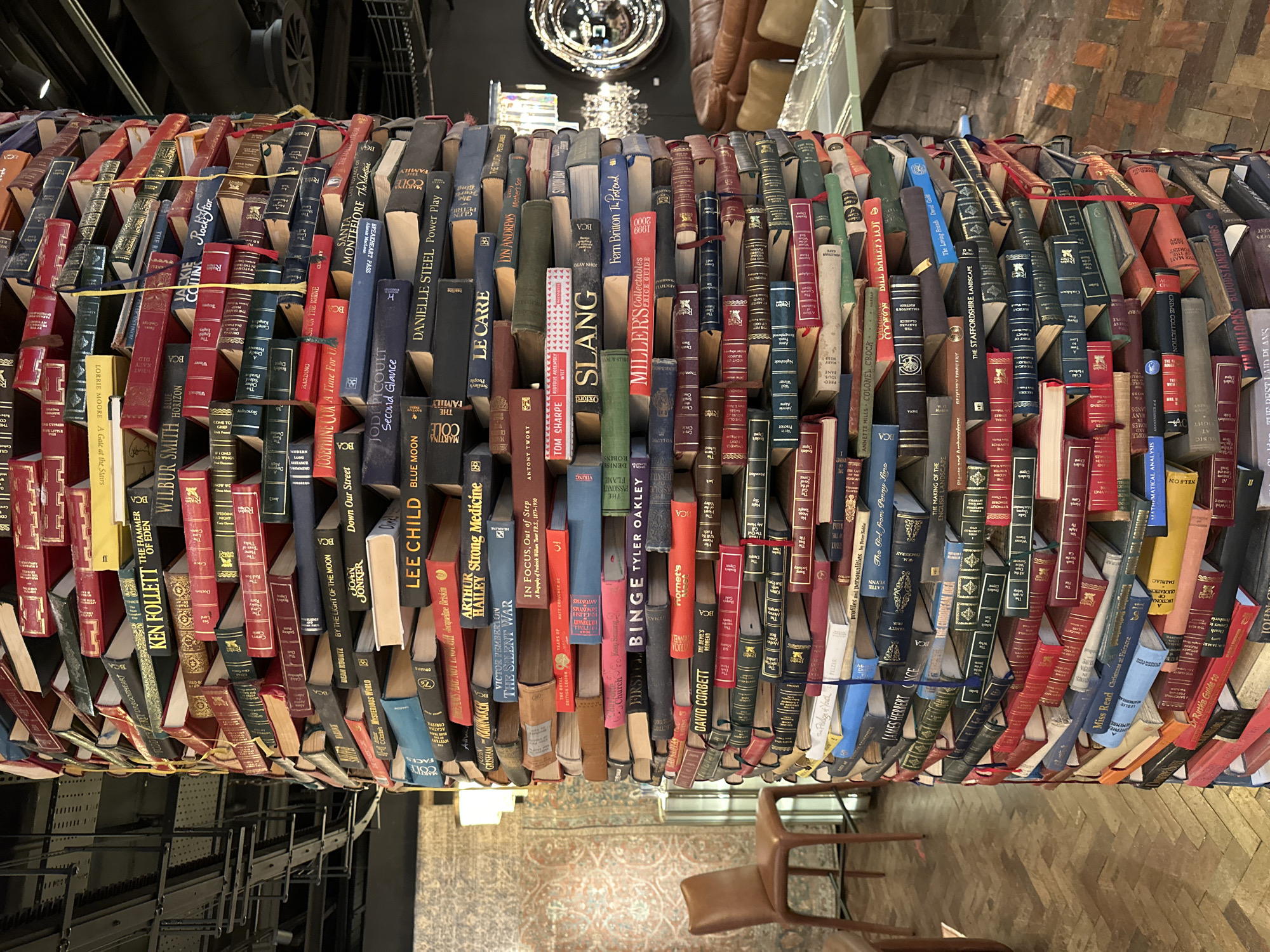 The image depicts a tall, cylindrical column constructed from an arrangement of books, situated indoors. The books are stacked horizontally and vertically, forming the structure's core, with spines facing outwards in many sections, revealing titles and authors. The titles visible suggest a collection of fiction and non-fiction works, including authors like Ken Follett, Wilbur Smith, Lee Child, Danielle Steel, and John Grisham.
The environment appears to be a well-lit interior space, possibly a retail establishment, library, or exhibition hall. The background includes furniture such as a brown leather sofa and chairs, a glass-topped table, and what appears to be a chandelier. The flooring is a parquet wood pattern. In the upper left portion of the image, industrial-looking metal structures are visible, suggesting a ceiling or mezzanine level. The lighting creates reflections on some of the book covers and the surrounding objects. No people are visible in the image.
While the image is described as being in London, United Kingdom, the specific location within London is not identifiable from the visual elements alone. The overall impression is one of artistic or architectural design utilizing books as a primary material.