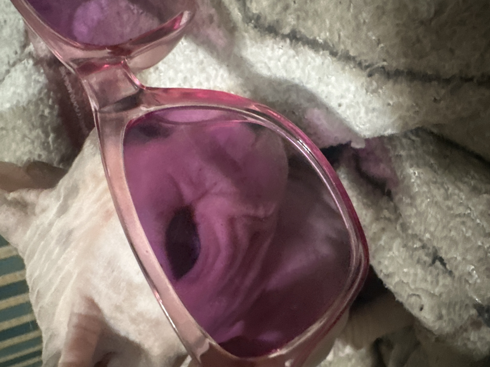 This media file features a close-up view of a pair of pink-tinted sunglasses resting on a textured, light-colored fabric, possibly a towel or blanket. The sunglasses have clear pink frames and lenses that tint the surrounding view with a purple hue. A reflection is visible within the right lens, showing a distorted image of what appears to be a person's face, with a dark circular area resembling an eye and lighter, curved lines suggesting facial features. The background fabric is soft and textured, with some darker fibers or shadows interspersed. The lighting appears to be artificial or from a diffused source, casting soft shadows. The image is cropped tightly on the sunglasses, with minimal background visible. No text is visible in the image. The location context of Togliatti, Russia, does not provide specific visual cues within the image itself. The overall impression is a still life focusing on the accessory and its reflective properties.