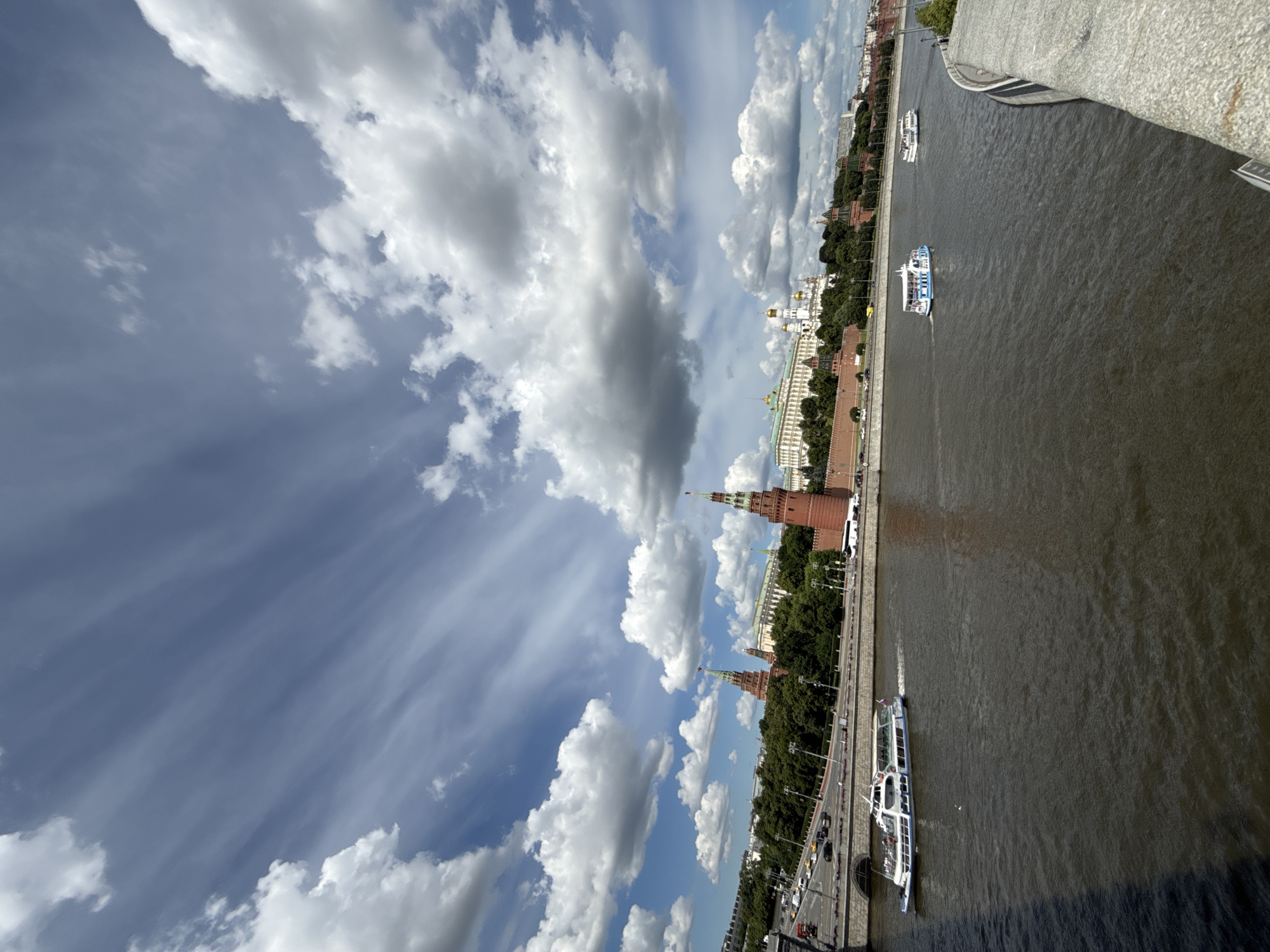 A wide-angle, high-angle shot captures a view of the Moscow Kremlin and the Moskva River on a partly cloudy day. The Kremlin's red brick walls and distinctive towers, including the Spasskaya Tower, dominate the mid-ground. Beyond the walls, the golden domes of cathedrals and white-washed buildings are visible. A line of lush green trees separates the Kremlin from the embankment.
The Moskva River flows in the foreground, a dark, rippling surface. Two passenger boats are navigating the river. The boat on the left is white with a blue stripe and appears to be a tour boat, with passengers visible on its deck. A second, smaller boat with a blue and white pattern is further downriver.
The embankment along the river features a road with traffic, including cars and buses. The sky above is a vibrant blue, streaked with wispy cirrus clouds and punctuated by larger, fluffy cumulus clouds, casting shadows on the landscape below. A stone bridge or embankment railing runs along the right edge of the frame, suggesting the vantage point is from a bridge or elevated walkway. The overall scene depicts a bustling urban landscape with historical landmarks under a dynamic sky.
