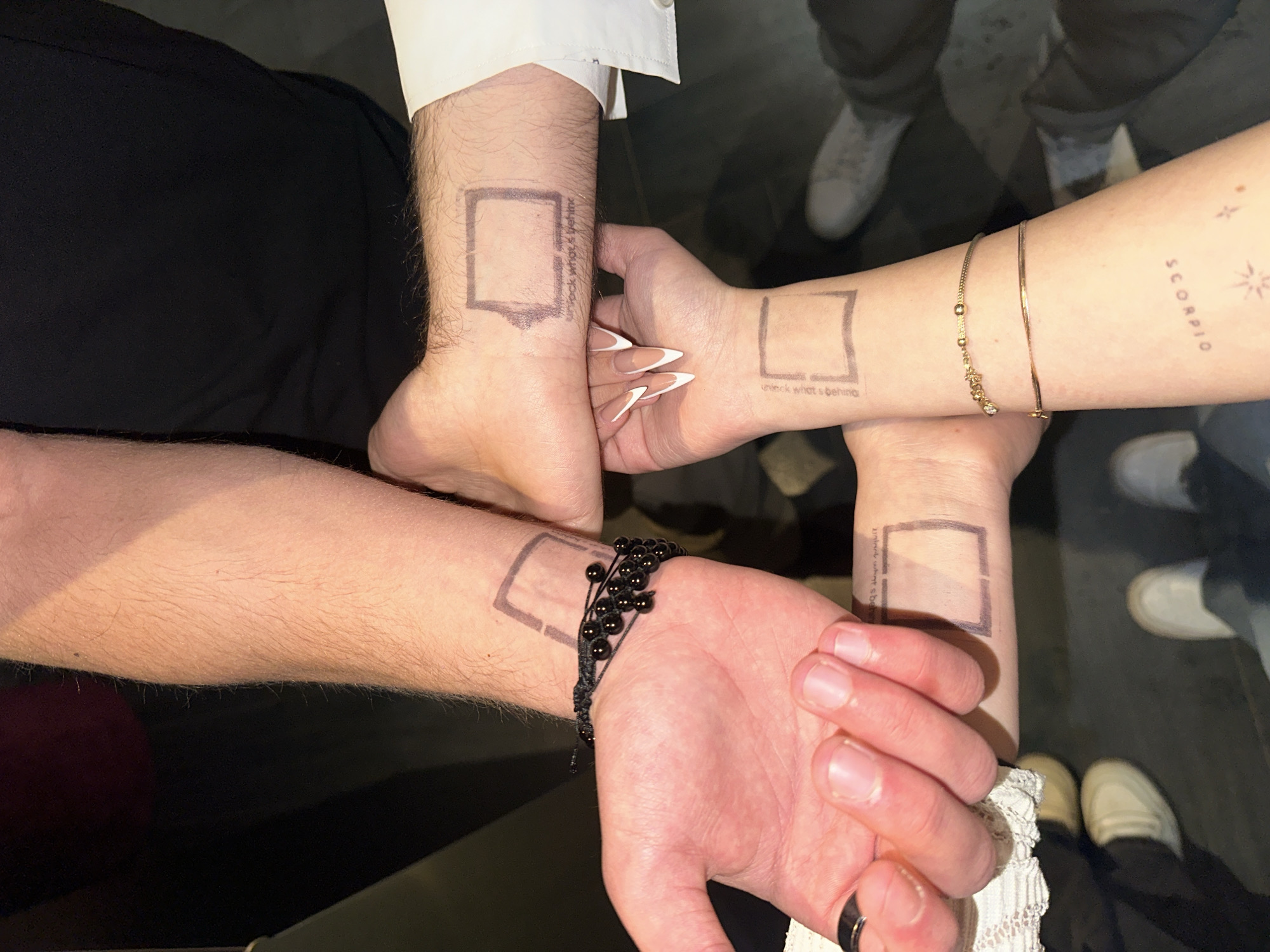 Four individuals' forearms and hands are partially visible in a close-up, top-down perspective, displaying their wrists. Each of the four wrists features a temporary mark: a stenciled, hollow rectangular outline, approximately 3-4 cm in length, with faint, blurred text beneath it that appears to read "unlock what's behind."
Essential details:
* **Subjects:** The image primarily focuses on four distinct human arms/wrists. One arm (upper left) is notably hairy and displays a black, multi-strand beaded bracelet. Another arm (upper right) is less hairy and shows a white dress shirt cuff. Two other arms (lower center and lower right) appear to be female; one hand has long, sharply pointed, white-tipped manicured nails, while the other arm (lower right) wears two slender gold-colored bracelets and has additional faint text "SCORPIO" and a star-like symbol tattooed or stamped further down the forearm. A black ring is visible on the index finger of the hand associated with the hairy arm.
* **Actions:** The subjects are holding their arms in close proximity, orienting their wrists towards the viewer, suggesting a collective display of the temporary marks.
* **Environment:** The background is dark and out of focus, indicating an indoor setting with ambient or low lighting. Blurred elements of clothing and footwear are visible, including dark fabric (possibly pants), a light-colored lace-trimmed garment, and multiple pairs of light-colored sneakers, consistent with a casual social gathering.
* **Location:** Based solely on the visual information provided in this image, a specific location within the city of Yerevan, Armenia, cannot be determined.