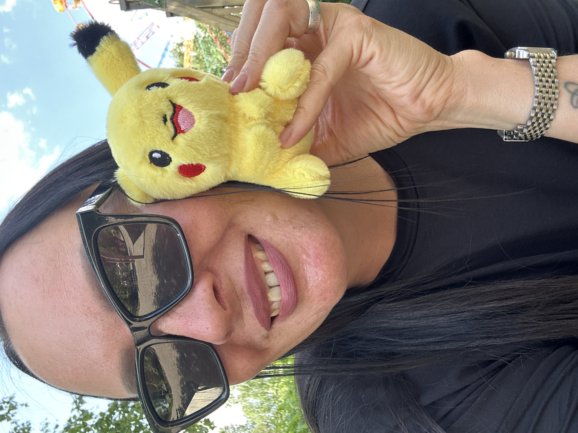 The image shows a woman with dark hair and sunglasses holding a Pikachu plush toy. She is wearing a black shirt and a watch. The background is blurry, but it appears to be outdoors, possibly in a park with some trees and a sky. The location is Unknown, Unknown.
