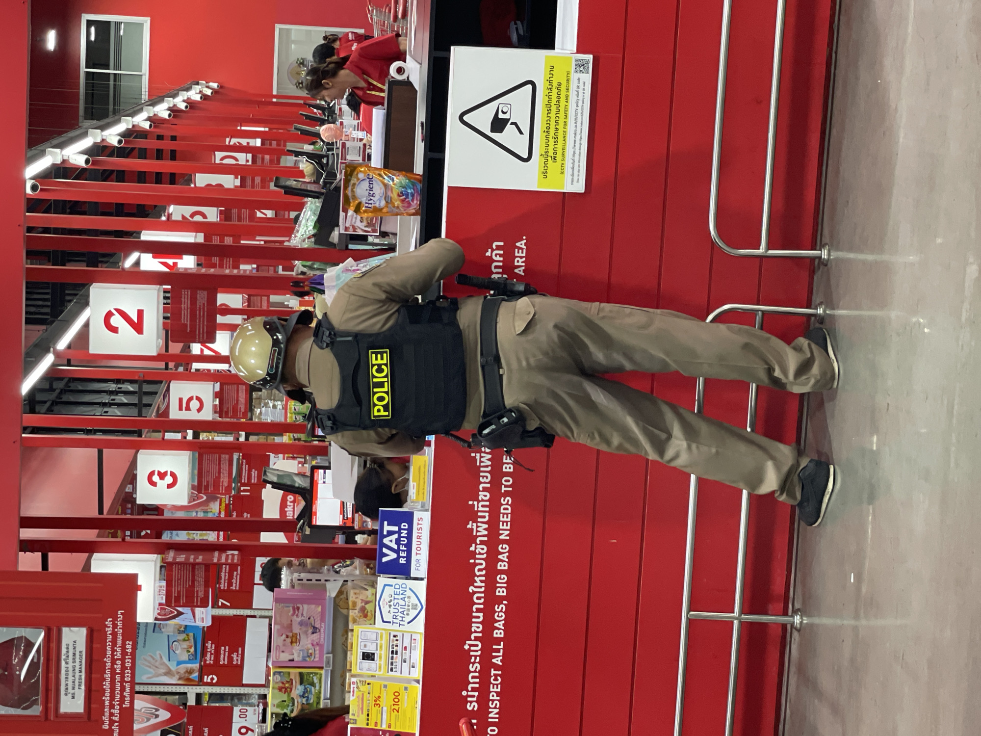 A male police officer, wearing a tan uniform, tactical vest with "POLICE" printed on the back, and a helmet, stands facing away from the camera in a store. The officer is positioned in front of a red counter, with shelves of products visible behind it. To the left of the officer, a sign in Thai and English reads, "DO TO INSPECT ALL BAGS, BIG BAG NEEDS TO BE..." Below this, a smaller sign displays a CCTV camera symbol and text in Thai and English, "CCTV SURVEILLANCE FOR SAFETY AND SECURITY." In the background, several checkout counters are visible, with other individuals, possibly store employees, attending to customers. The environment appears to be a retail store, likely in a commercial area. The scene is indoors, with bright lighting. The overall impression is one of security presence within a shopping environment.