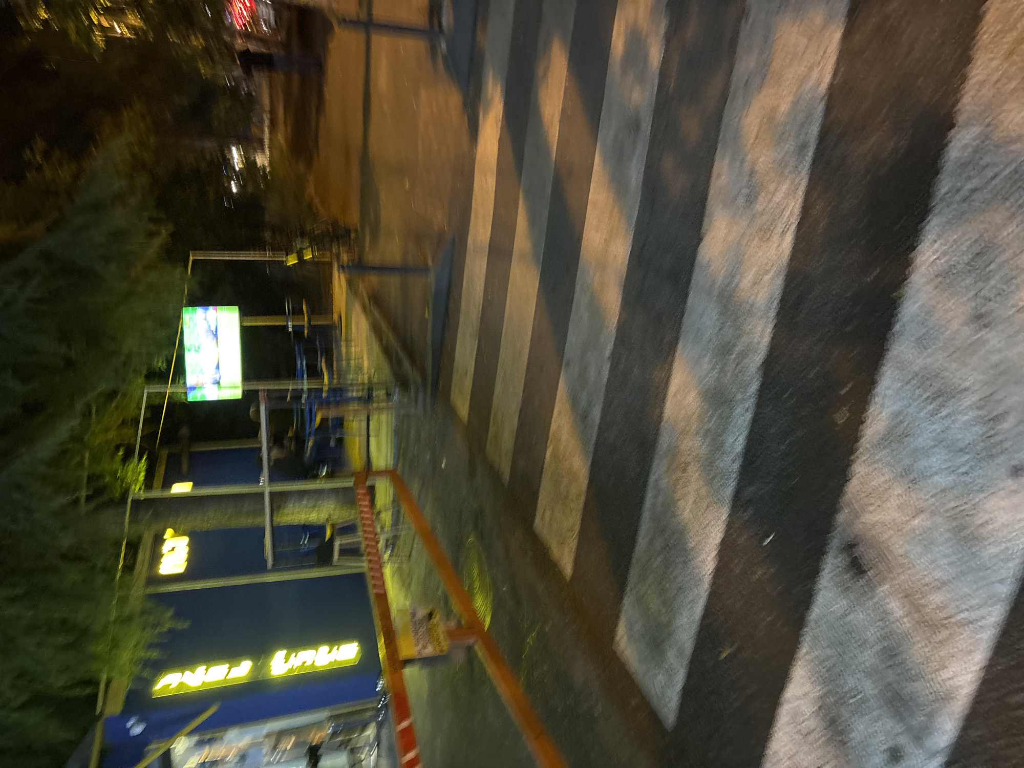 A nighttime street scene in Tbilisi, Georgia, shows a pedestrian crossing with striped white and grey markings. An orange barrier with reflective tape is positioned across the pedestrian crossing. In the background, a building with blue siding features bright yellow neon lettering that reads "RUSTAVI 2". Adjacent to the building, a tree trunk is visible. A television screen displays a colorful image. Further into the background, a shadowy figure stands near a street with car lights visible. The overall lighting suggests an urban environment at night.