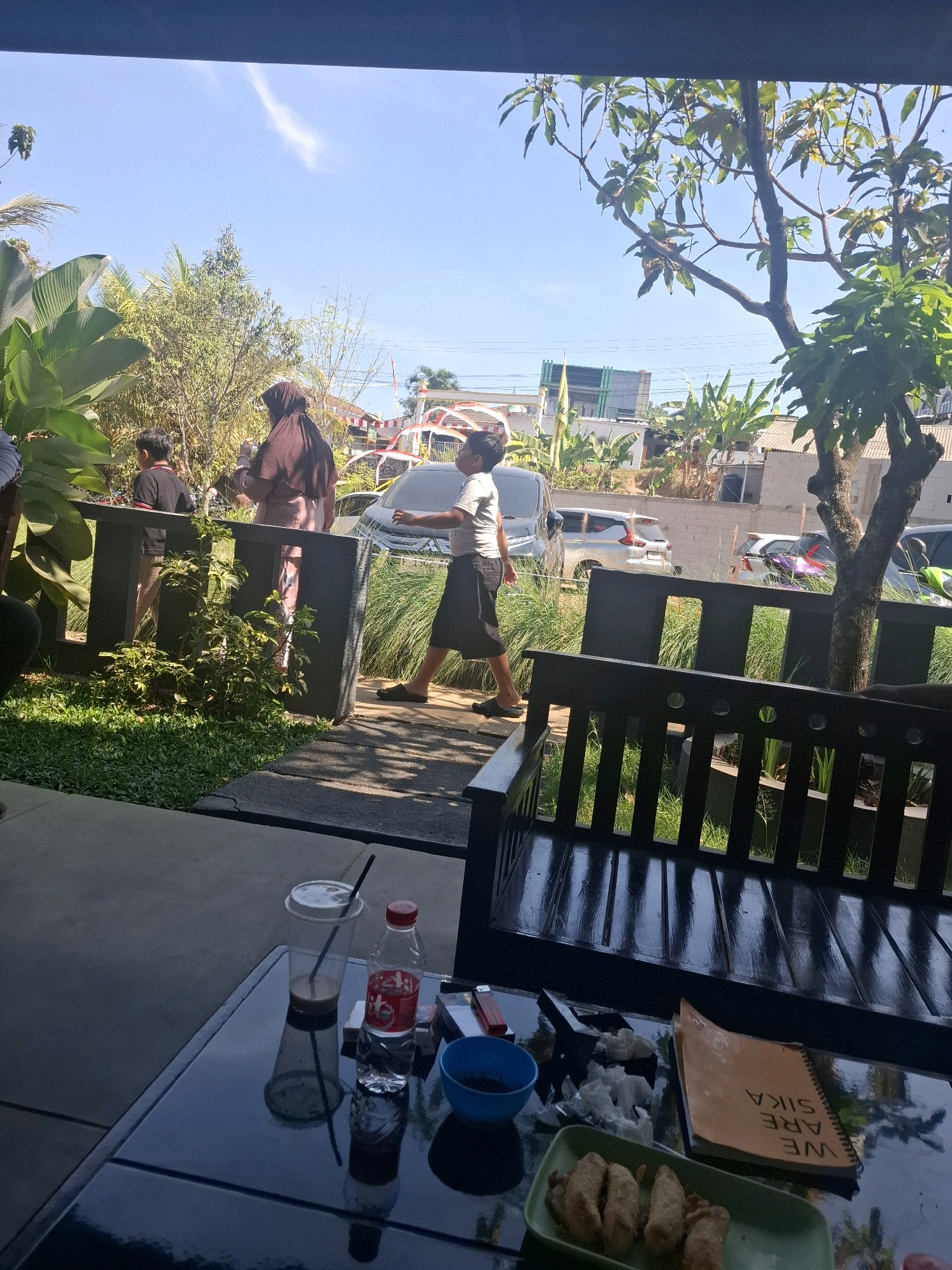 The image depicts a scene outdoors, likely in Cibentang, Indonesia, with a focus on a table in the foreground and people in the background. On the glossy black table, a clear plastic cup with a dark liquid and a straw, a bottle of water with a red cap and label, a lighter, tissues, a small blue bowl, a green rectangular plate with several fried food items, and a spiral-bound notebook with the words "WE ARE SIKA" printed on its orange cover are visible.
In the mid-ground, several individuals are present. A person wearing a brown hijab and a long dress walks past a dark fence. A young boy in a dark shirt and shorts is seen walking away from the viewer, looking down. Another person in a light-colored t-shirt and dark shorts walks in the same direction. Behind them, cars are parked.
The environment includes lush green vegetation, trees with broad leaves and thinner branches, and a bright blue sky with a wispy white cloud. Buildings with tiled roofs and some modern structures are visible in the distance. The scene suggests an outdoor seating area, possibly a cafe or restaurant, with a view of a street or parking area. Sunlight casts shadows on the ground and the table, indicating a bright, sunny day.