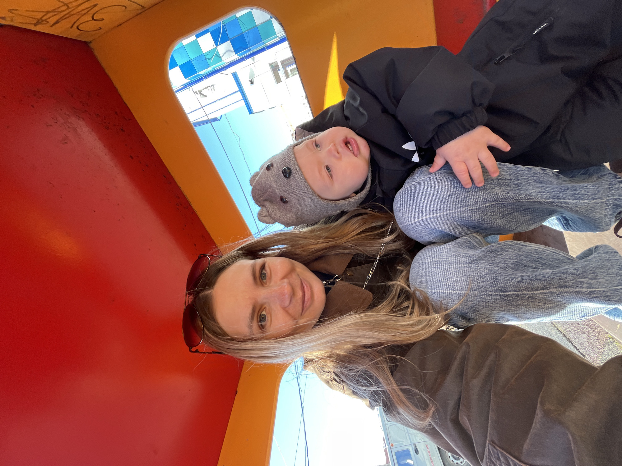 A selfie taken in Chaikovsky, Russia, shows a woman and a baby inside a playground structure. The woman is fair-skinned with long blonde hair and is wearing sunglasses on her head, a brown jacket, and jeans. She is smiling and looking at the camera. The baby, who is fair-skinned and wearing a gray hat and a black winter jacket, is sitting on her lap. The playground structure has red, orange, and yellow surfaces. Outside the structure, the sky is visible, and there are buildings with a blue-and-white checkered pattern. A white van is also visible in the background.