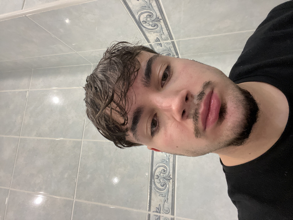 A close-up portrait depicts a male individual against a tiled wall, likely indoors in Vila Real, Portugal. The subject has wet, dark brown, disheveled hair, brown eyes, a mustache, and a chin beard. His gaze is directed slightly downwards, and his lips appear full and slightly pouted. He is wearing a black crew-neck t-shirt. The background wall is composed of light blue-grey rectangular ceramic tiles, some exhibiting reflections from light sources. A horizontal decorative border tile, featuring a blue and white intricate pattern, is visible behind the subject's head and shoulders.