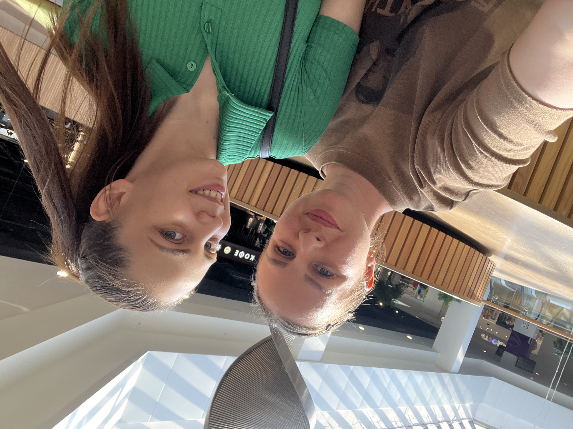 Two women are in a selfie taken indoors. The woman on the left has long brown hair and wears a green ribbed top. The woman on the right has blonde hair pulled back and wears a brown long-sleeved shirt. A partial view of the interior features modern architecture with white walls and a wood-paneled area in the background. A cafe sign reading "КОФЕ" is visible. The location is in Izhevsk, Russia.