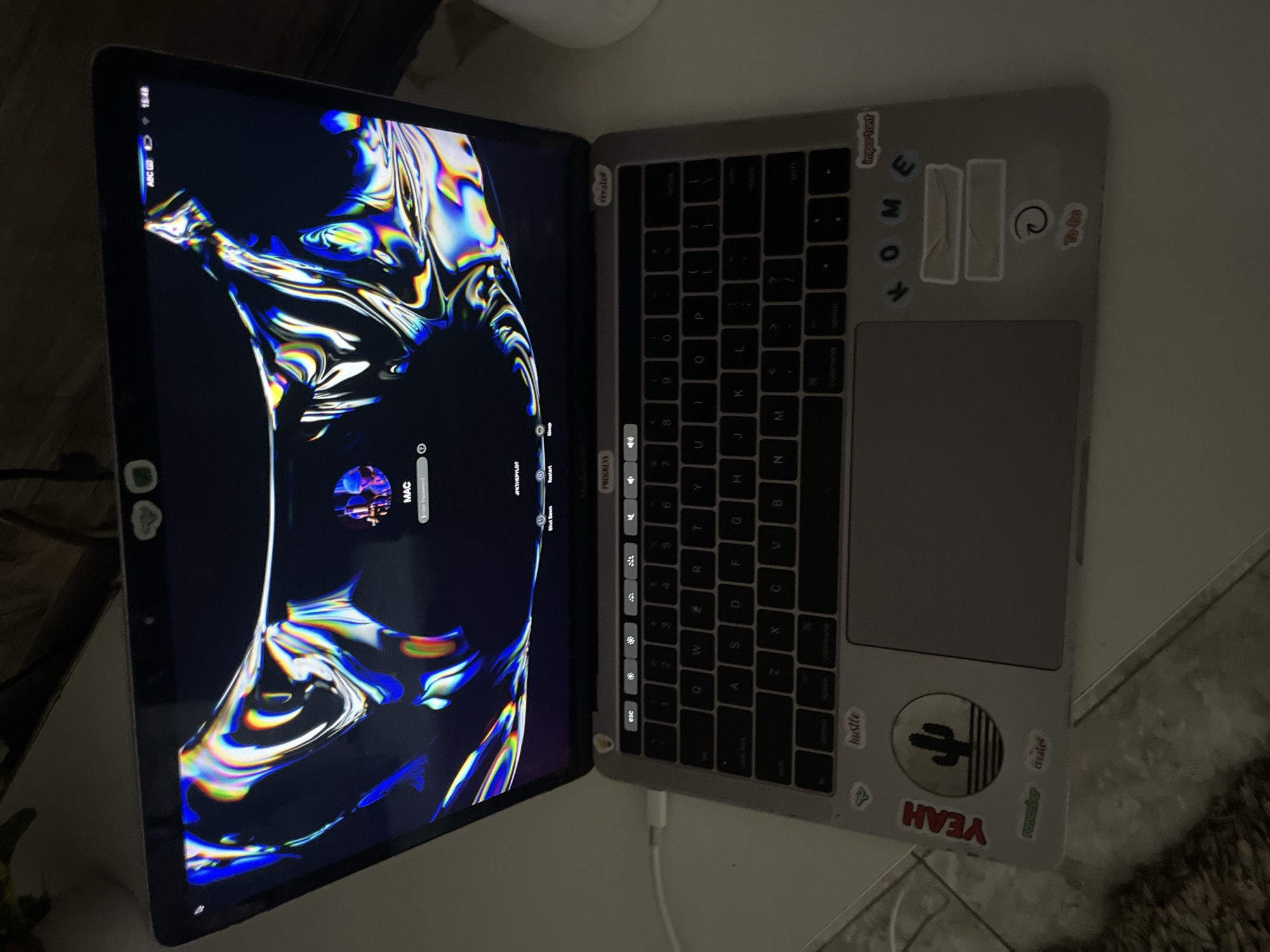 An open MacBook laptop rests on a light-colored surface, likely a desk, in an indoor setting in Kuchingoro, Nigeria. The laptop's screen displays a login interface at 3:48 PM, featuring a vibrant, abstract wallpaper with iridescent, flowing colors. The user account "MAC" is shown, prompting for a password, with options to "Shut Down" and "Restart" visible. The top left corner of the screen shows "15:48", "ABC", and icons for battery and Wi-Fi.
The laptop's palm rest and area around the keyboard are adorned with numerous stickers. Visible text on these stickers includes "YEAH", "hustle", "remember", "create", "important", and individual letter stickers forming "HOME". A prominent circular sticker depicts a cactus and a sun, while another sticker reads "To-Do". Other partially visible stickers include "PROGRESS" and "JFKTHEPYLOT". A white charging cable is connected to the laptop's left side. No individuals are present in the frame, and the room appears dimly lit, with the screen providing the main illumination. The activity is minimal, with the laptop awaiting user input to log in.