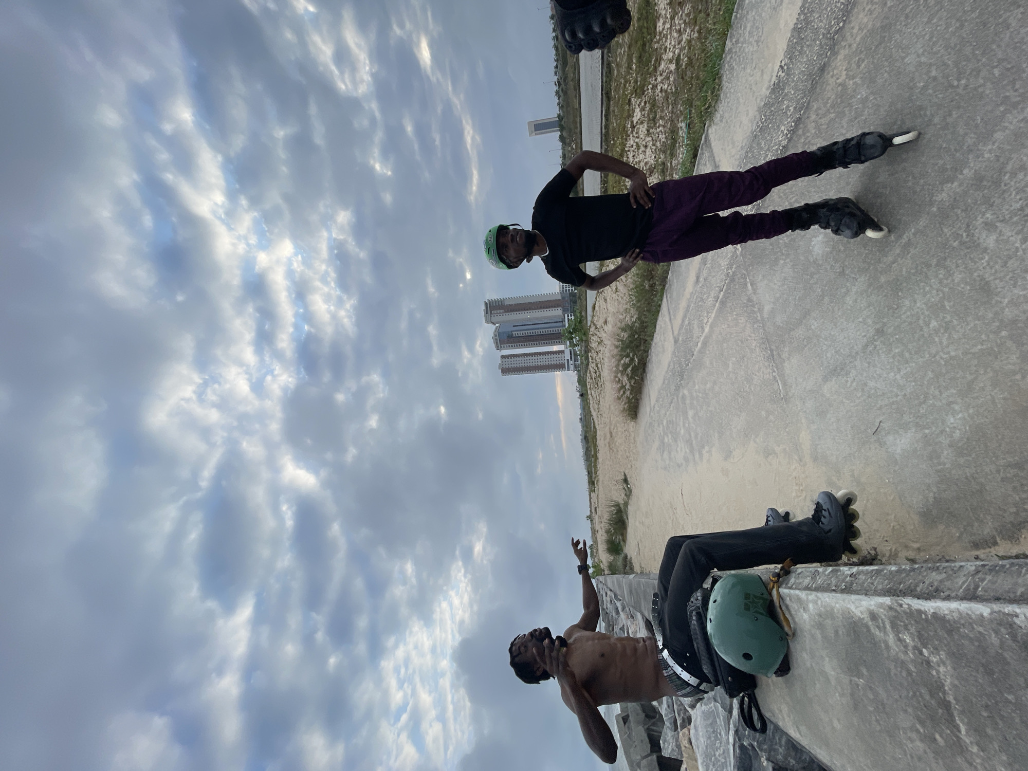 The image depicts a coastal scene with a cloudy sky, possibly at dusk. In the foreground, two men are present. On the left, a shirtless man with dark skin and braided hair is seated on a rocky embankment, wearing dark pants and a checkered belt. He is gesturing with his hands and appears to be talking. A green helmet and a dark bag are placed next to him. On the right, a man wearing a black t-shirt, purple pants, and a green helmet is standing on rollerblades. He has his hands on his hips and is looking upwards. In the background, there is a sandy area with sparse vegetation, a paved walkway, and several modern high-rise buildings in the distance, suggesting an urban or developing area. The location is identified as Ilado, Nigeria.