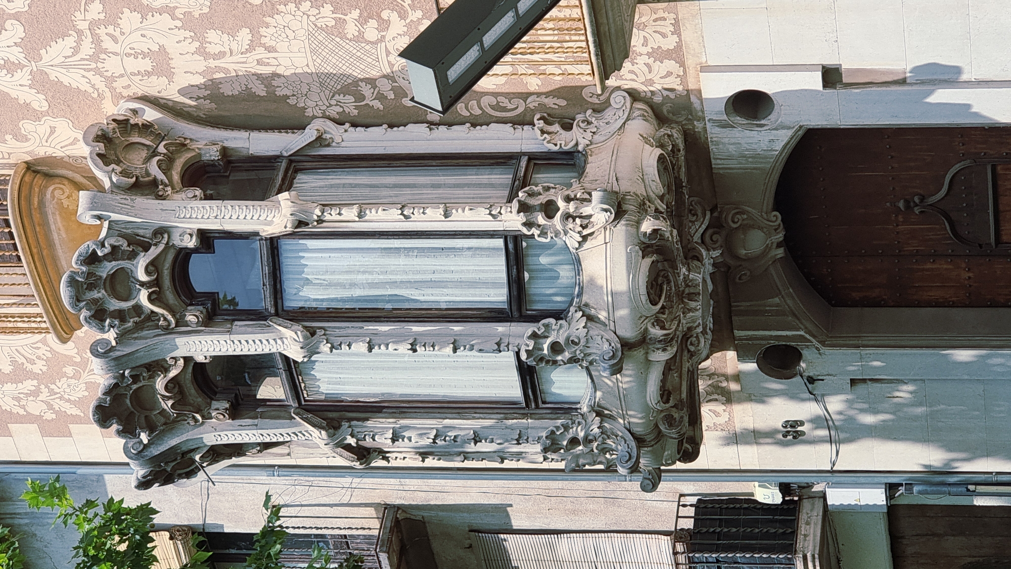 The image shows a close-up of a highly ornate, stone balcony on the facade of a building. The balcony features intricate carvings resembling organic forms, possibly flora, with a central bay window framed by elaborate sculptural elements. The window itself has dark framing and is covered by sheer, white curtains. Above the bay window, the building's facade is decorated with a repeating floral pattern in shades of pink and beige. A modern, angular light fixture is mounted to the right of the balcony, casting a shadow. Below the balcony, an arched wooden door with studded detailing is visible, along with the building number "38" and two circular openings in the stone wall. The setting appears to be a European city, possibly Olot, Spain, given the architectural style which is reminiscent of Art Nouveau or Modernisme. The lighting suggests it is daytime, with strong sunlight creating distinct shadows. There are no people visible in the frame.