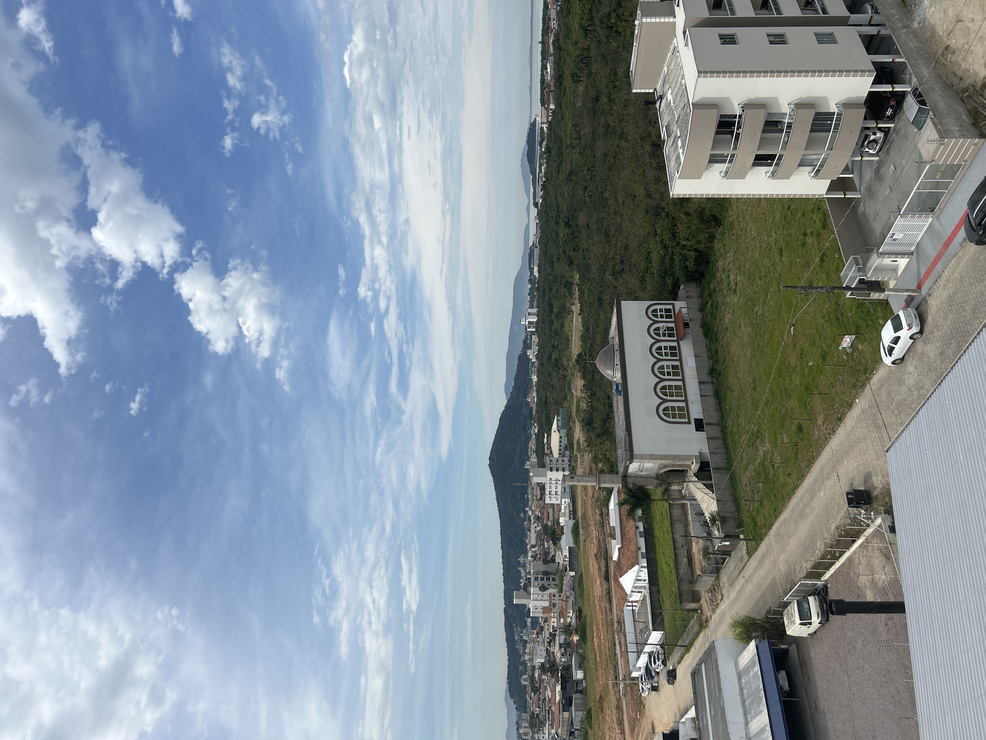 The image is an aerial view of a coastal city in Palhoça, Brazil, under a partly cloudy sky. In the foreground, a modern apartment building with beige and white facades and balconies faces a street. To the left, a light-colored van is parked next to a building with a blue awning. Across the street, a white building with a minaret and arched windows, resembling a mosque, is visible. Further in the distance, a sprawling urban landscape with residential and commercial buildings stretches towards a large, green, tree-covered mountain. The background features a hazy body of water, likely the Atlantic Ocean, with islands and mountains in the far distance. The overall scene depicts a blend of urban development and natural scenery in a Brazilian coastal town.