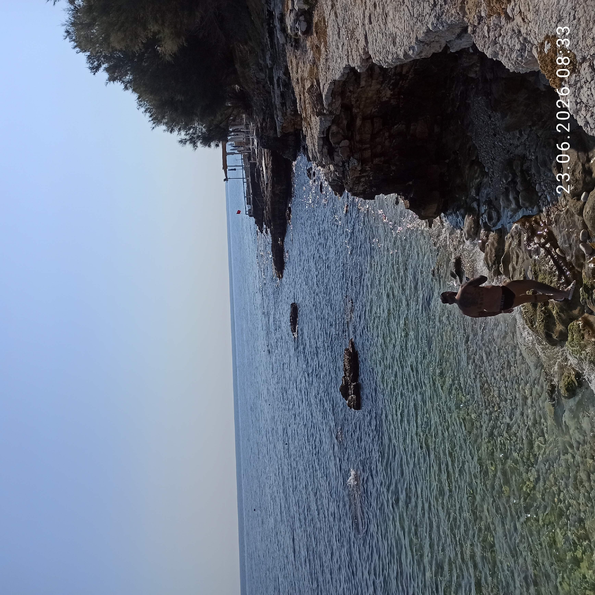 A man with tattoos walks along a rocky shore on a clear, sunny day. The man is wearing black swimming trunks and white sandals. He is walking towards the water, which is clear and shallow near the shore, with visible pebbles. The shoreline is rocky with some larger rocks and sparse vegetation. In the background, the sea stretches to the horizon under a clear blue sky. There is a small wooden structure with awnings and chairs visible on the rocky outcrop to the right, along with a red flag. The timestamp "23.06.2026 08:33" is visible in the bottom right corner of the image. The scene depicts a serene coastal environment, possibly in Molfetta, Italy, given the timestamp's date format.
