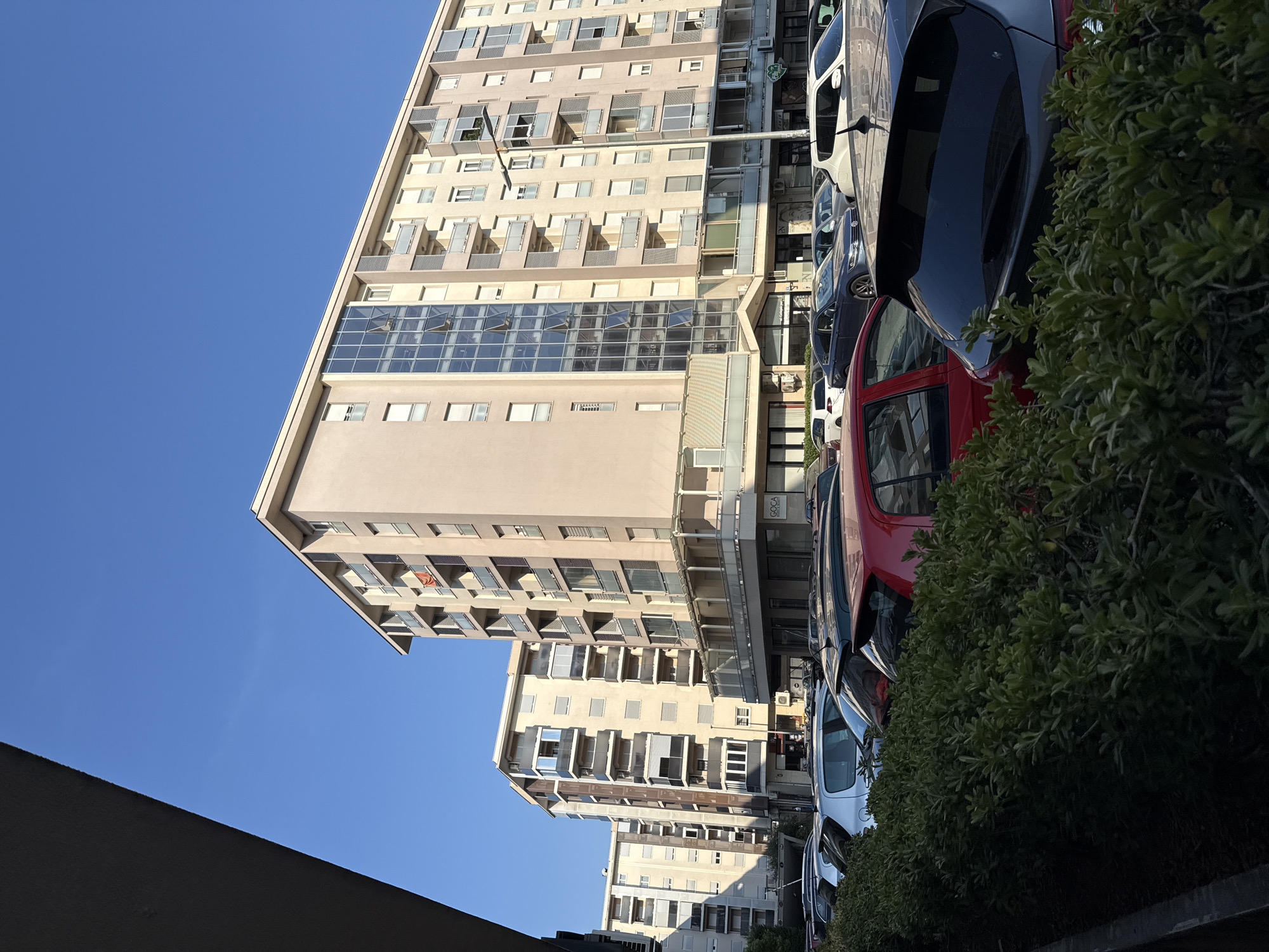 In a city setting, a row of parked cars is visible in front of several residential buildings under a clear blue sky. A lush green hedge runs along the foreground, partially obscuring the cars. The buildings are multi-story apartment complexes with numerous balconies and windows, indicative of an urban residential area. The sunlight casts shadows on the ground, suggesting it is daytime. There are no people visible in the frame. The cars are of various colors and models, parked in a lot or on a street. The scene appears to be a typical urban environment.