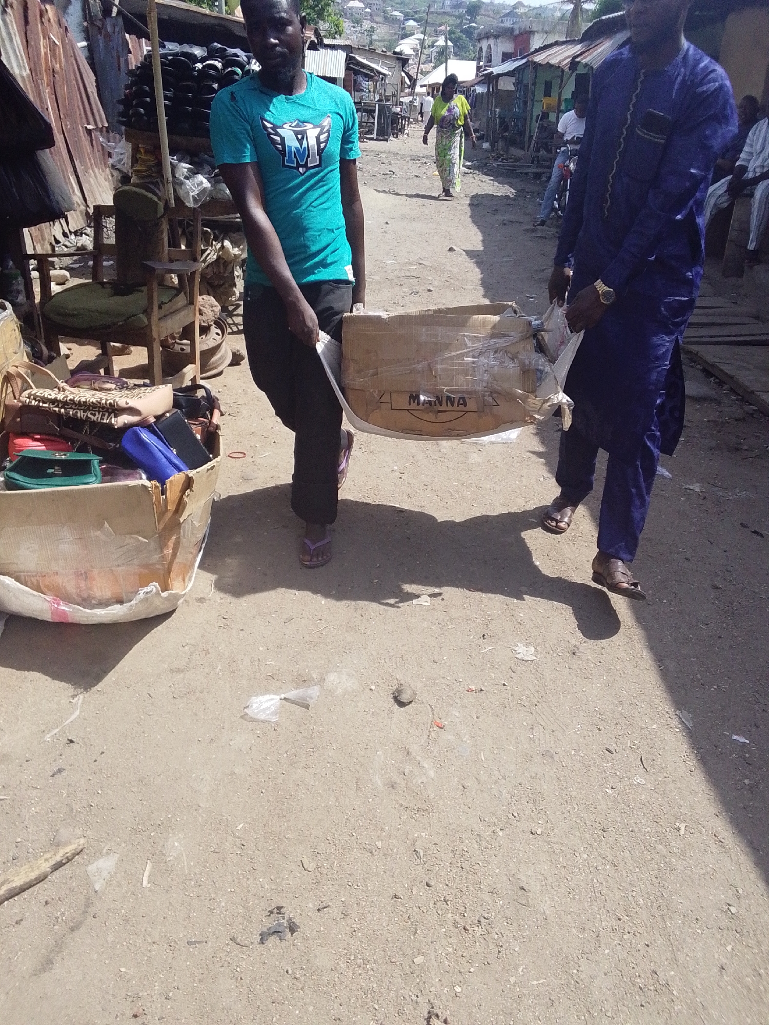 This media file captures a vibrant outdoor street scene in Lokoja, Nigeria, under bright, clear sunlight, casting distinct shadows on the dirt and gravel ground.
In the foreground, two men are actively engaged in carrying a large, light-colored cardboard box down the road. The man on the left wears a teal t-shirt with a blue and white winged "M" logo, black trousers, and purple flip-flops. The man on the right is dressed in a traditional dark blue long-sleeved outfit with gold embroidery and brown sandals. The cardboard box they are carrying has the word "MANNA" printed on its side.
To the immediate left, a makeshift market stall is visible, featuring a wooden chair with a green cushion and what appears to be a display of footwear on a stand. Another open cardboard box in the lower left corner is filled with assorted handbags and purses, one of which clearly displays the brand name "VERSACE."
In the mid-ground and background, the street is lined with simple structures, many topped with corrugated iron roofs, indicating market stalls or shops. A woman in a patterned green and white dress walks further down the road. Other individuals and a partially visible motorbike suggest a busy local environment. The scene depicts a typical day of commerce and movement in a bustling community setting.