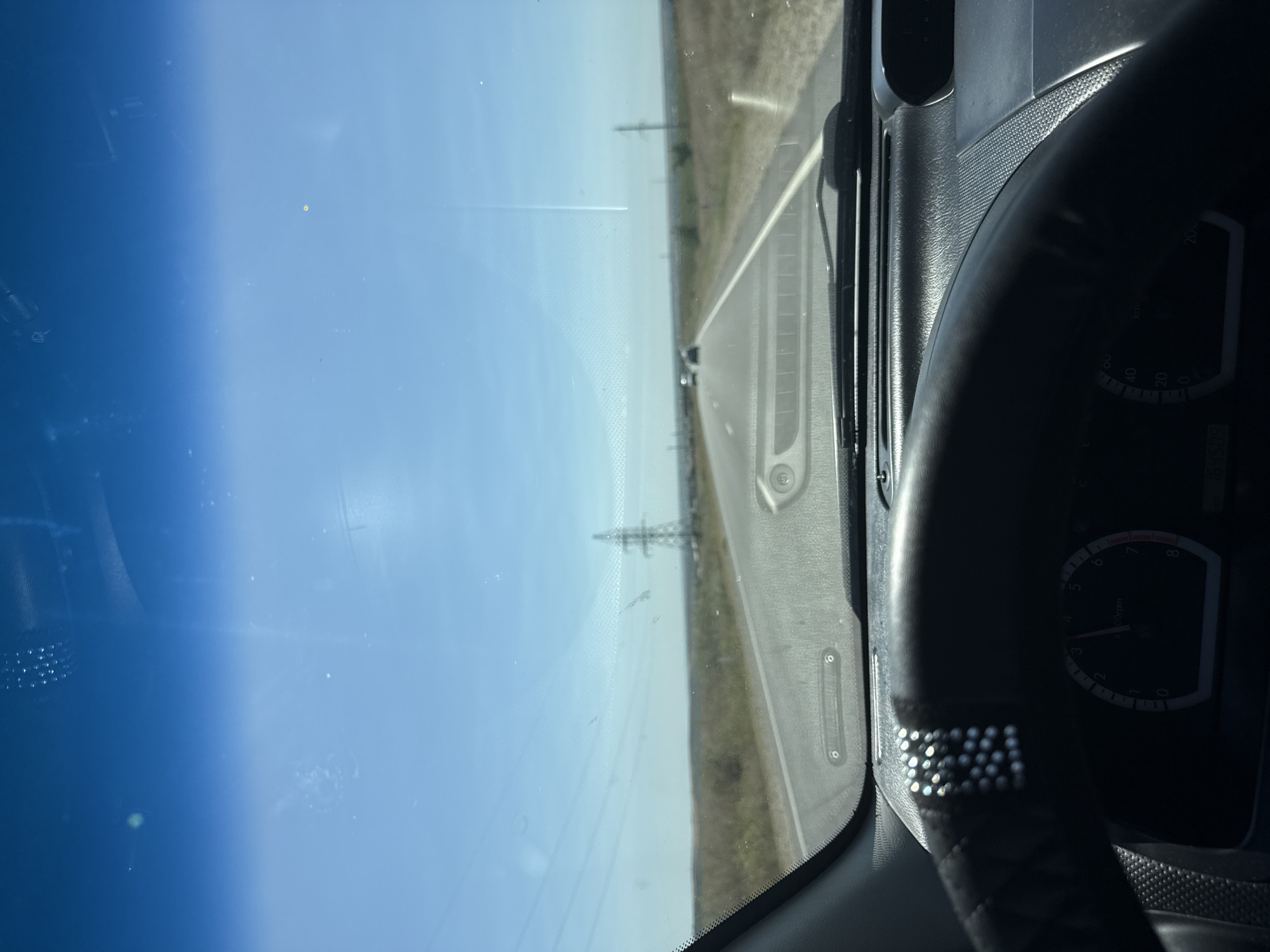 The image is a first-person view from the driver's seat of a car traveling on a two-lane asphalt road. The road stretches into the distance under a clear blue sky, with dry, grassy plains on either side. In the background, power lines and a tall transmission tower are visible. The dashboard and steering wheel are in the foreground. The steering wheel is black and appears to have a decorative strip of small, reflective beads on its upper left side. The speedometer shows the needle indicating a speed between 0 and 2,000 rpm. The scene is set on a sunny day. The location is the road leading to Cioc-Maidan, Moldova.
