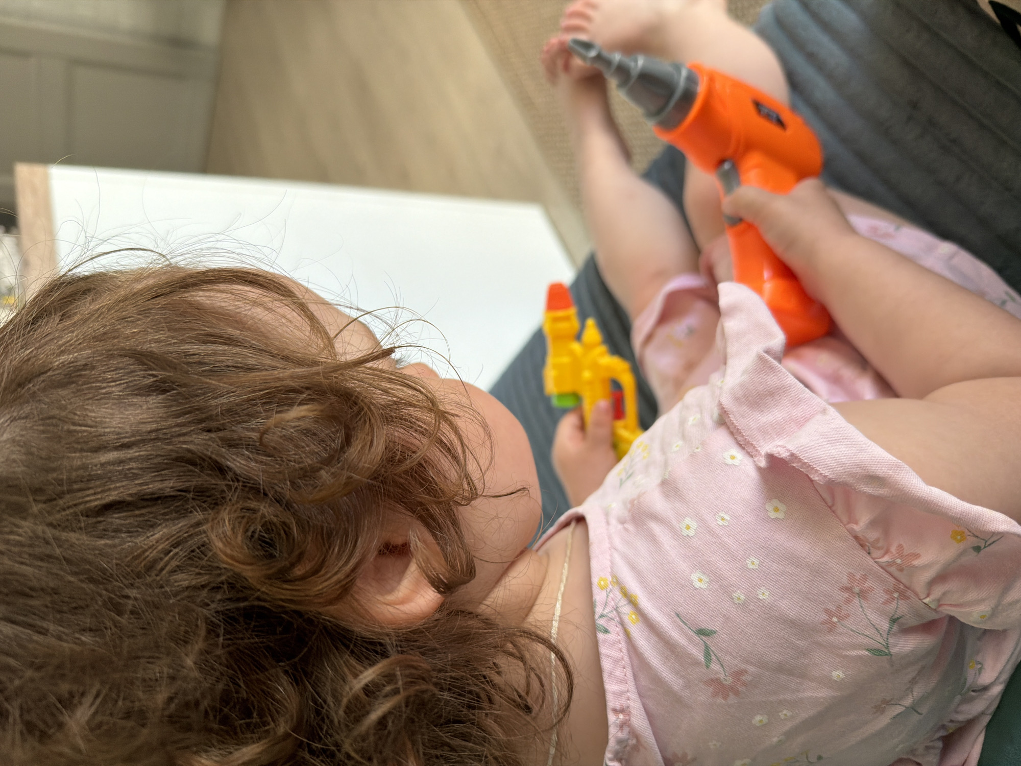 A child with curly brown hair is lying down, holding a yellow toy gun in one hand and an orange toy drill in the other. The child is wearing a pink floral patterned shirt. The location is not specified, so it cannot be determined if the image was taken in the city of Pervomaysky, Russia.