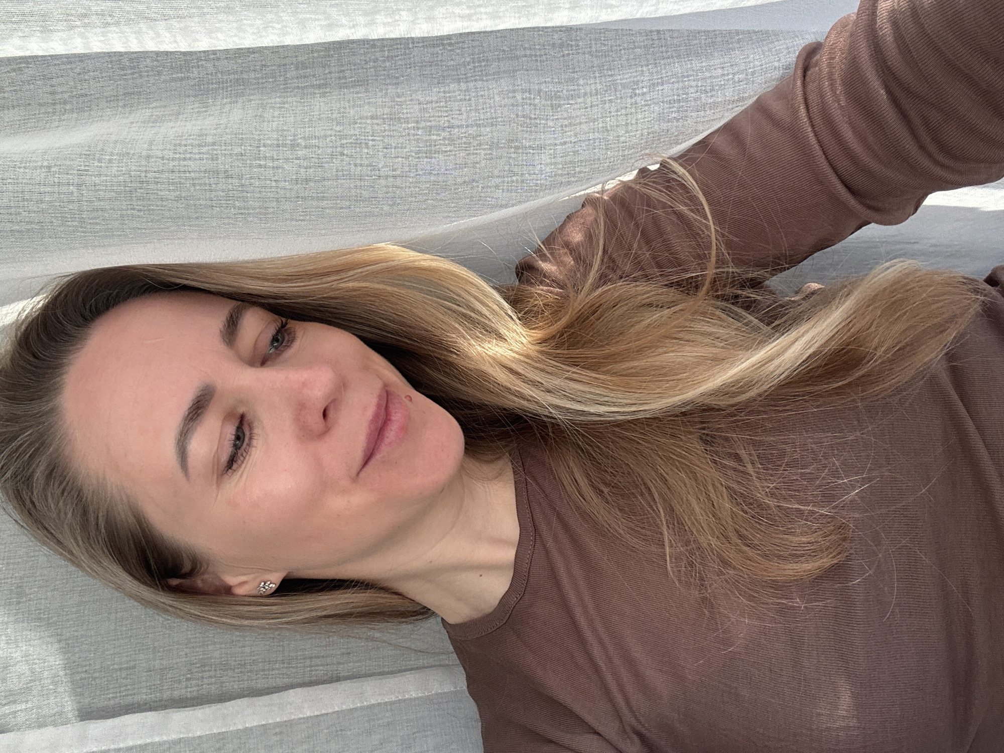 A fair-skinned woman with long blonde hair is lying down, possibly on a bed. She is wearing a brown long-sleeved shirt. Part of her face is visible, showing her left eye and cheek. A white curtain drapes above her head. The image was likely taken indoors. There is no evidence to suggest the location is Chaikovsky, Russia.