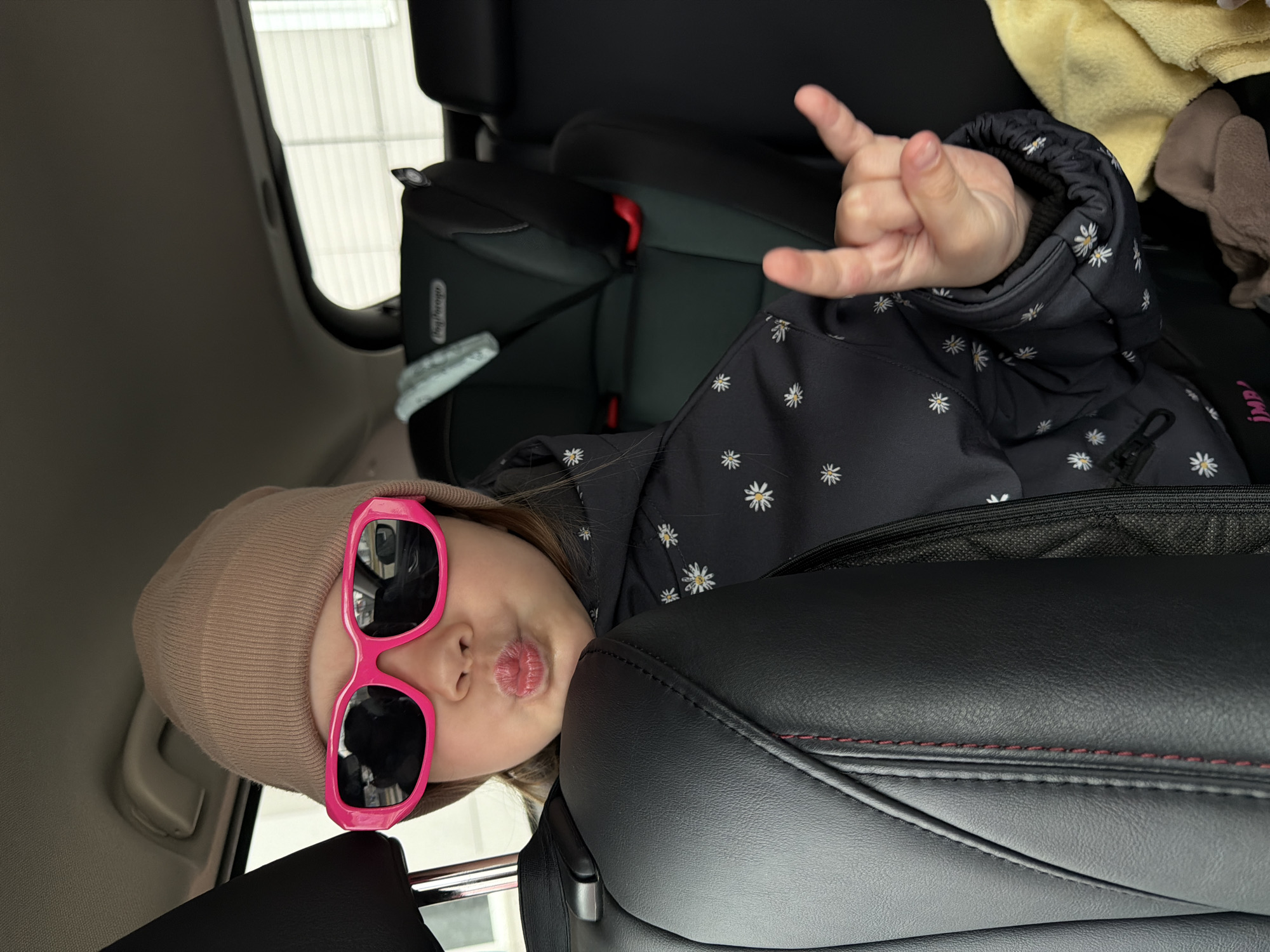 A young child is playfully posing in the back seat of a car in Chaikovsky, Russia. The child, wearing a beige beanie and large pink sunglasses, is dressed in a dark jacket patterned with white daisies. Her head is tilted, and her lips are pursed in a "kissy" or "pouty" expression. One hand is raised, making a "rock on" gesture.
The child is positioned in the back seat, seemingly looking over the top of a black leather front seat, which features red stitching. In the background, another child car seat, identifiable by the "Joie" brand logo, is visible. Through the car window, an exterior building with horizontal paneling can be seen, indicating it is daytime. A yellow towel or blanket and other soft belongings are stowed in the area behind the front seats. The child's jacket also has a small pink tag that reads "iMP!". The attire suggests cooler weather. The overall scene conveys a cheerful and mischievous mood.