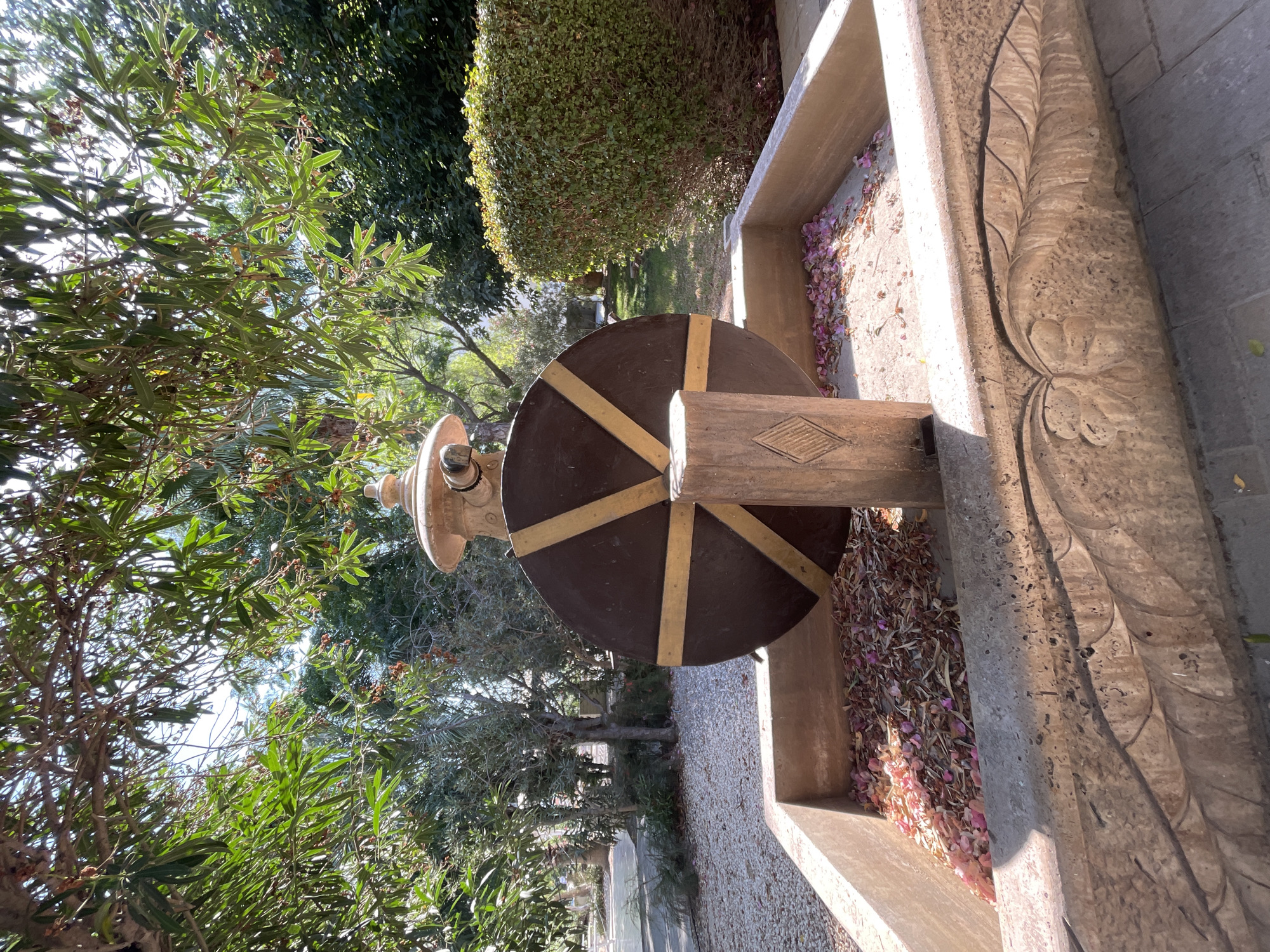 An outdoor decorative water feature is depicted, featuring a large, dark brown, circular wheel with golden spokes, positioned behind a carved wooden post within a rectangular stone basin. The basin contains dried leaves and pink petals. Behind the wheel, a stone structure with a rounded top is visible. The scene is set amidst lush greenery, with trees and a trimmed hedge in the background. Sunlight filters through the leaves, casting shadows. The foreground shows the carved details of the stone basin's edge, resembling leaves. The location is identified as Trikomo, Northern Cyprus / Cyprus.