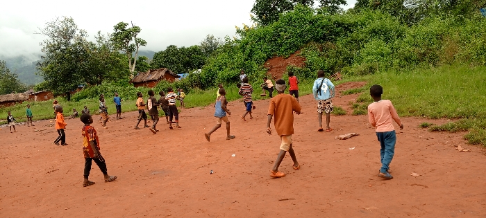 The scene depicts approximately twenty children, predominantly male, engaged in outdoor activity on a reddish-brown dirt clearing in the city of Ngalula, Democratic Republic of the Congo. The children, ranging in apparent age from approximately five to fifteen years old, are spread across the foreground and midground. Many are in various states of motion, some running or walking, suggesting a game or informal play. Their attire consists of diverse casual clothing, including t-shirts, shorts, and pants; some wear sandals or shoes, while others are barefoot.
In the background, several traditional structures with earthen walls and reddish-brown or thatched roofs are visible amidst scattered green grass and trees. A dense, verdant hill covered with lush vegetation forms the backdrop on the right side of the image, while on the left, distant mountains shrouded in haze are visible under an overcast sky. The ground of the clearing is uneven, with some scattered debris, possibly small stones or fragments of wood. No specific play equipment or delineated boundaries are present. The overall environment suggests a rural or semi-rural community setting with significant natural elements.