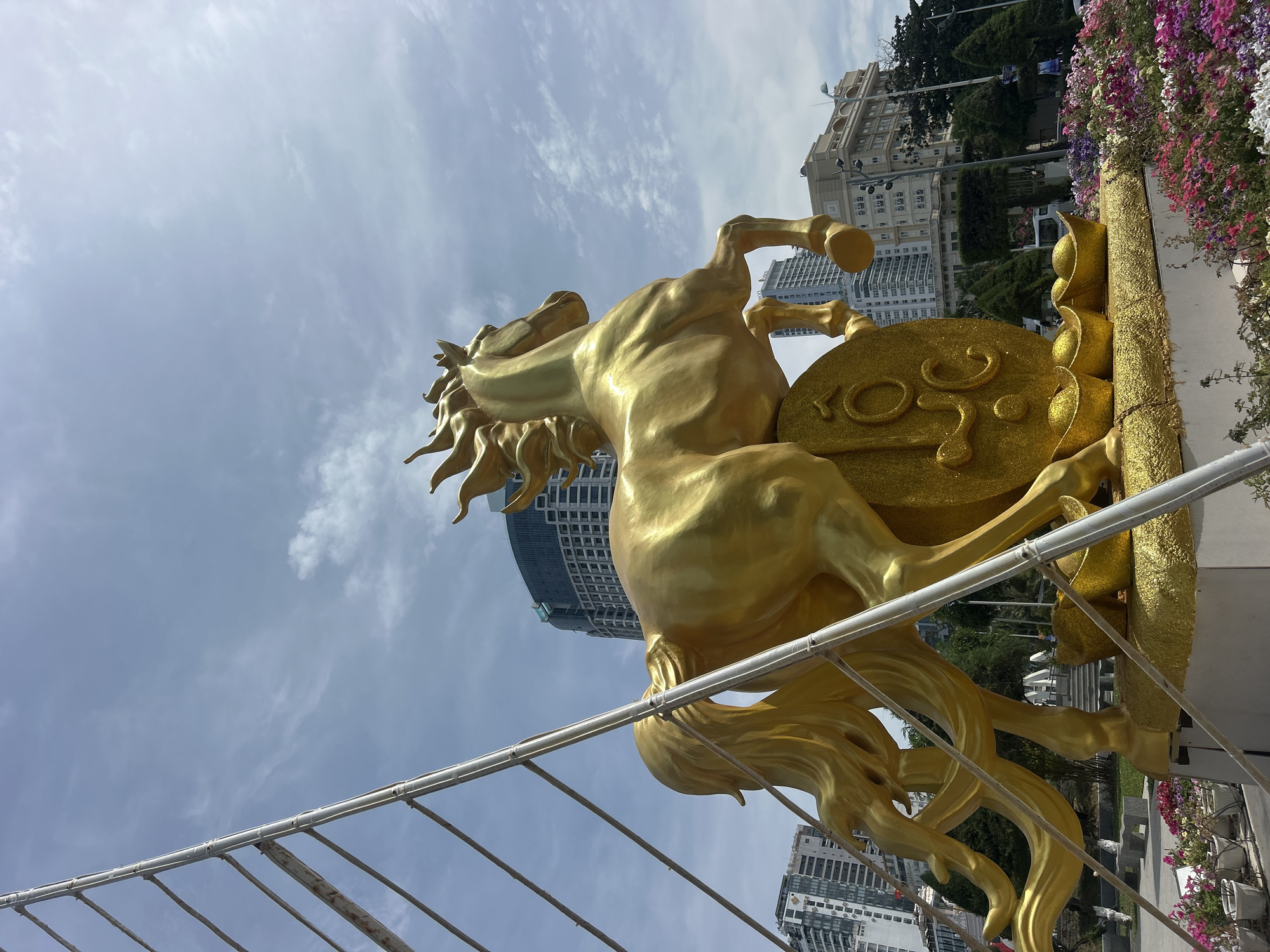 A low-angle, outdoor, medium shot depicts a gold horse statue in the city of Nha Trang Ward, Vietnam. The statue is rearing up, with its front legs raised, and it is positioned in front of a large gold coin-shaped object. The background includes several buildings and a cloudy sky. A metal structure or fence is visible in the lower part of the frame. The location is likely a public space or a tourist attraction within the city. The statue is the main focal point, and its gold color contrasts with the surrounding environment.