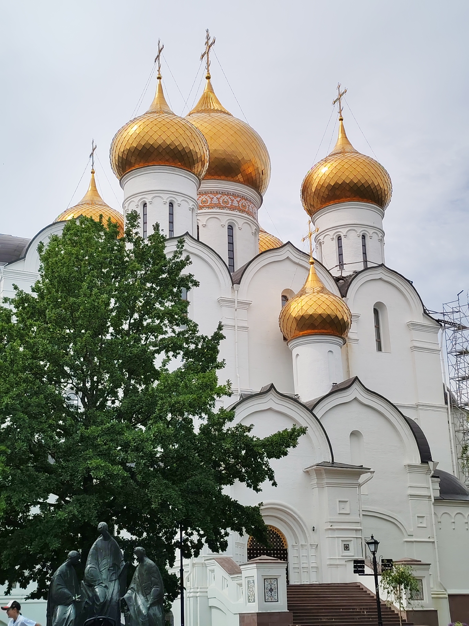 In Rusanovo, Russia, a large white cathedral with multiple golden domes topped with crosses is the central subject. A large green tree with dense foliage partially obscures the left side of the cathedral. In the foreground, a bronze sculpture depicts three figures wearing robes. To the right of the cathedral, scaffolding is visible. The sky is overcast. A person wearing a white shirt and a red and white cap is partially visible on the far left, near the bottom of the frame. A lamppost stands on the right side of the steps leading to the cathedral entrance.