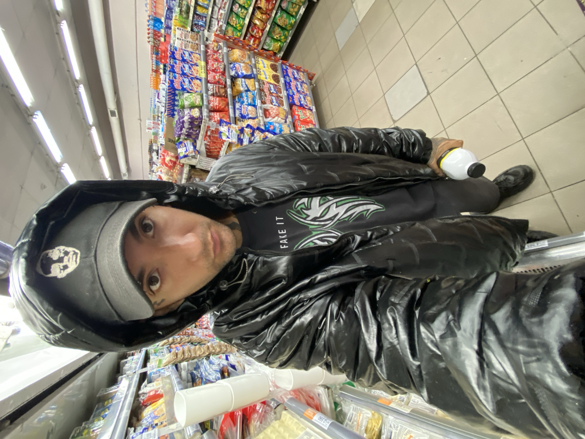 A person is standing in a supermarket aisle in Buenos Aires, Argentina, taking a selfie. They are wearing a black, shiny hooded jacket, a gray baseball cap with a white logo, and a black t-shirt with a design. The person is holding a white and black bottle in their left hand. Shelves of snacks, including chips and candies, are visible behind them. A refrigerated section with various products is in the foreground. The floor is tiled in light gray and white. Fluorescent lights illuminate the scene.