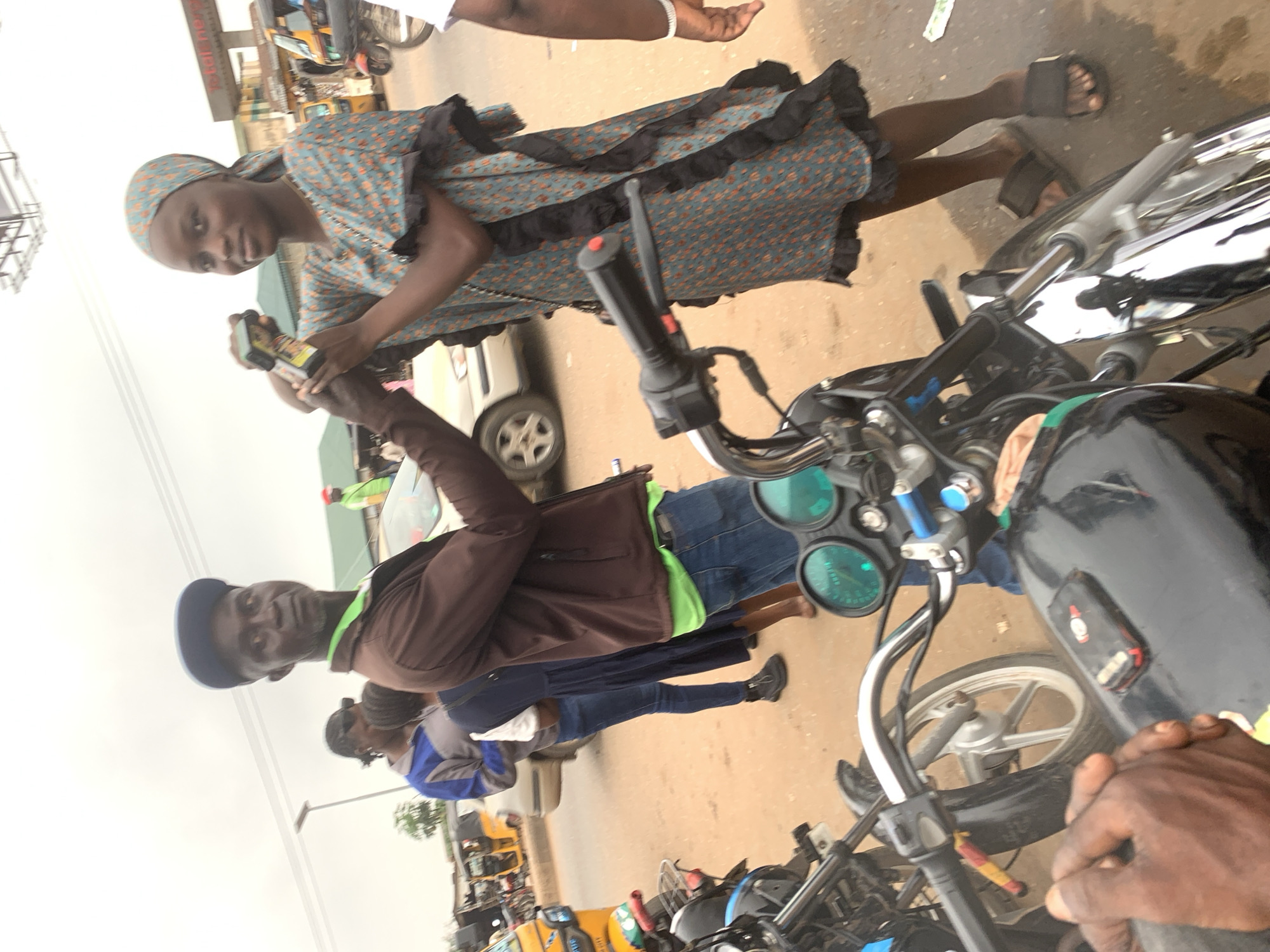 In Shagamu, Nigeria, a man wearing a blue cap and a brown jacket over a green shirt stands with his left arm extended towards a woman. The woman, wearing a patterned headwrap and a blue and orange dress with black ruffled trim, holds a small object, possibly a phone or a small device, in her outstretched hands. Her right hand is near the object. A motorcycle is in the foreground, with its handlebars, wheel, and dashboard visible. Another motorcycle is partially visible to the left. In the background, other individuals, vehicles including a yellow tricycle, and buildings are present. The ground is sandy and dusty. The sky is overcast.